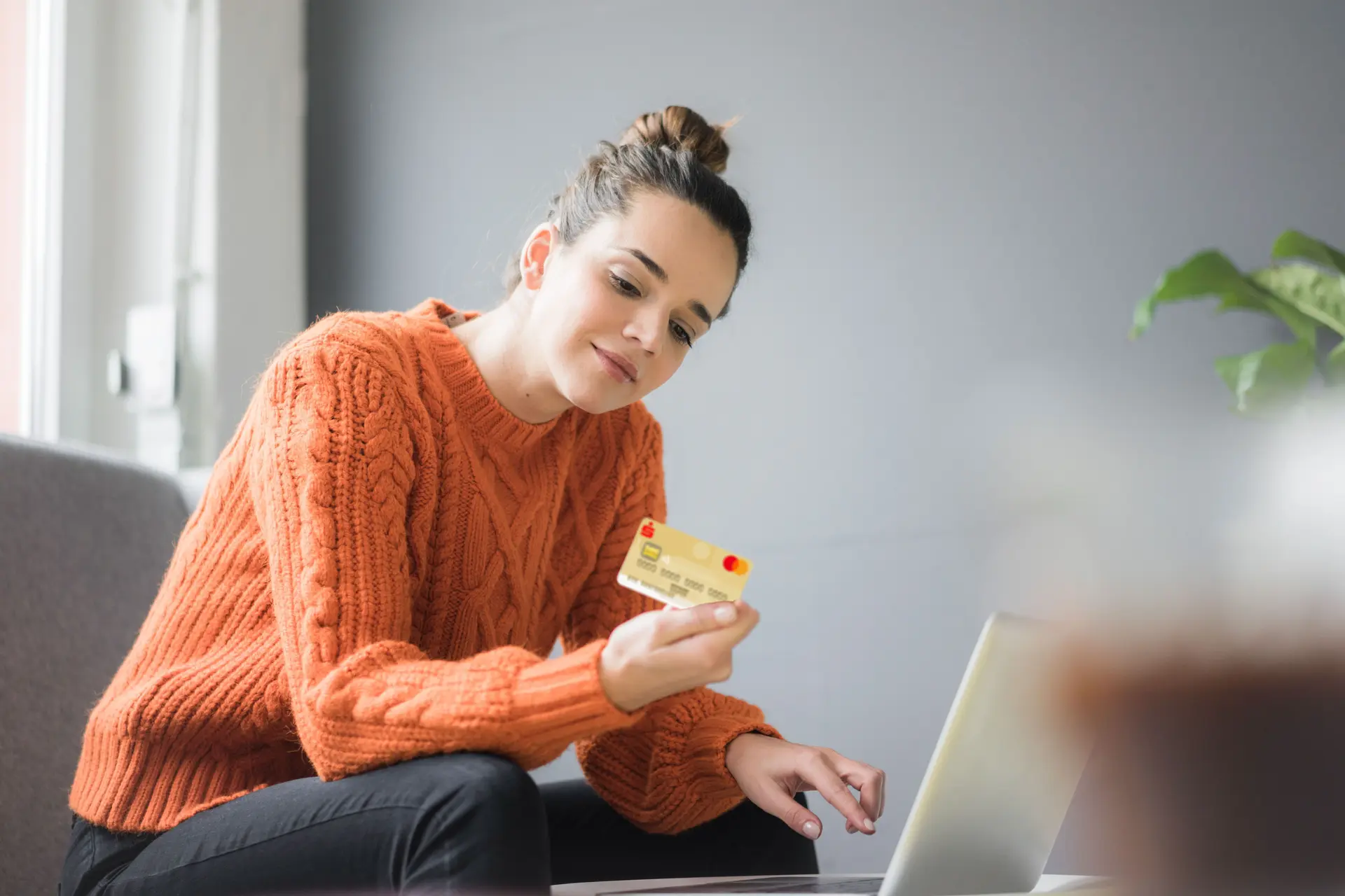 Eine junge Frau in orangenen Pulli sitzt vor einem Laptop. In der Hand hat sie eine goldene Kreditkarte. Sie schaut auf die Rückseite der Kreditkarte.
