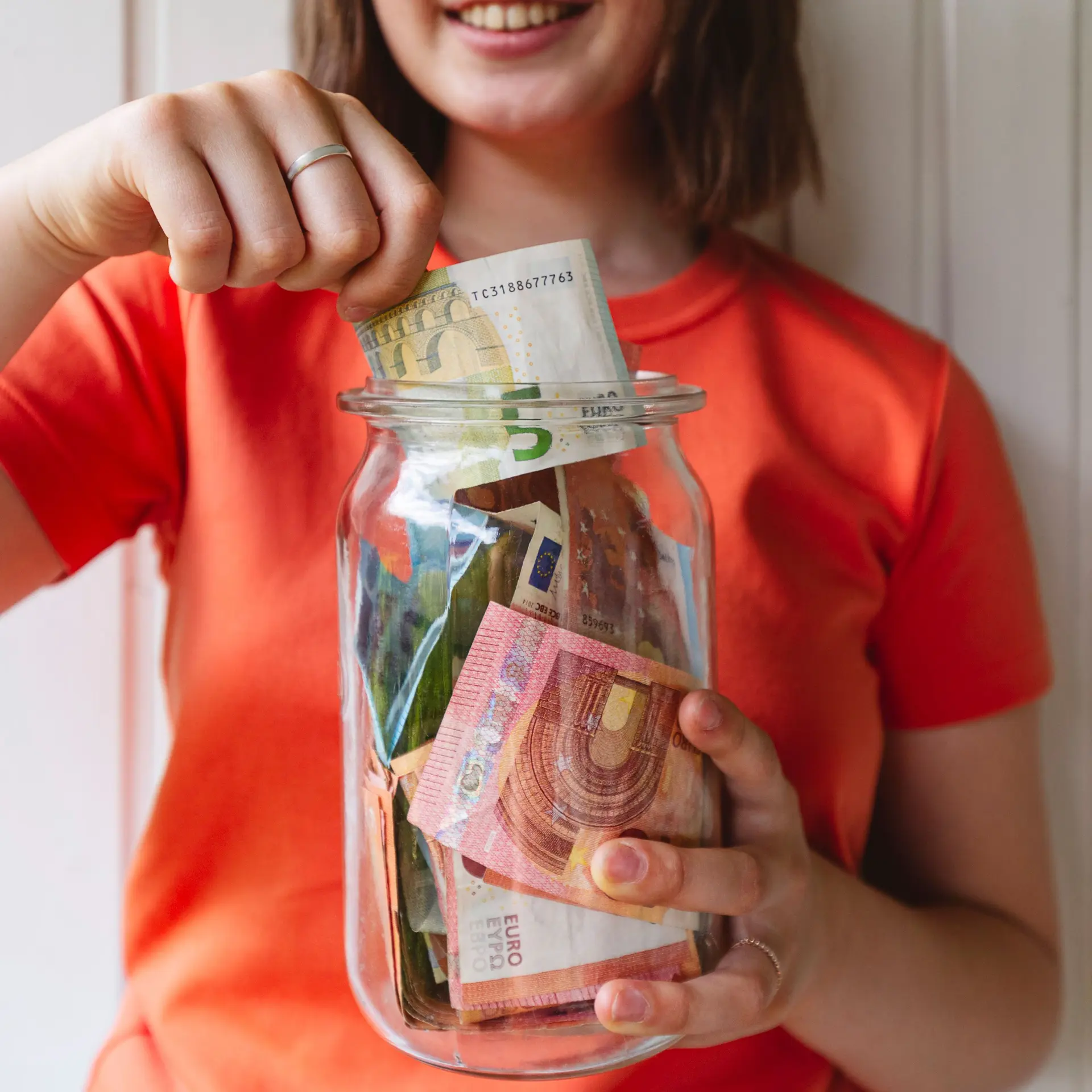 Oberkörper einer jungen Frau mit einem Glas voller Geld vor sich. mit einer Hand steckt sie Geld ins Glas.