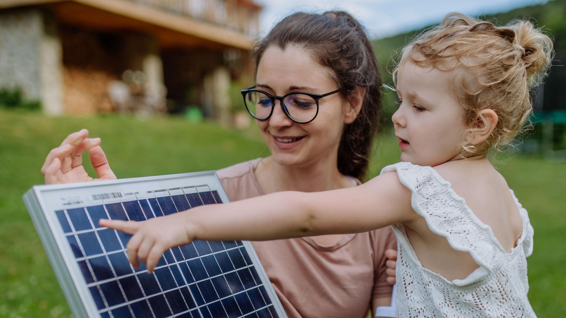 Eine junge Frau zeigt ihrer Tochter ein Solarpanel. Sie stehen auf einer Wiese. Im Hintergrund ist ein Haus zu sehen.