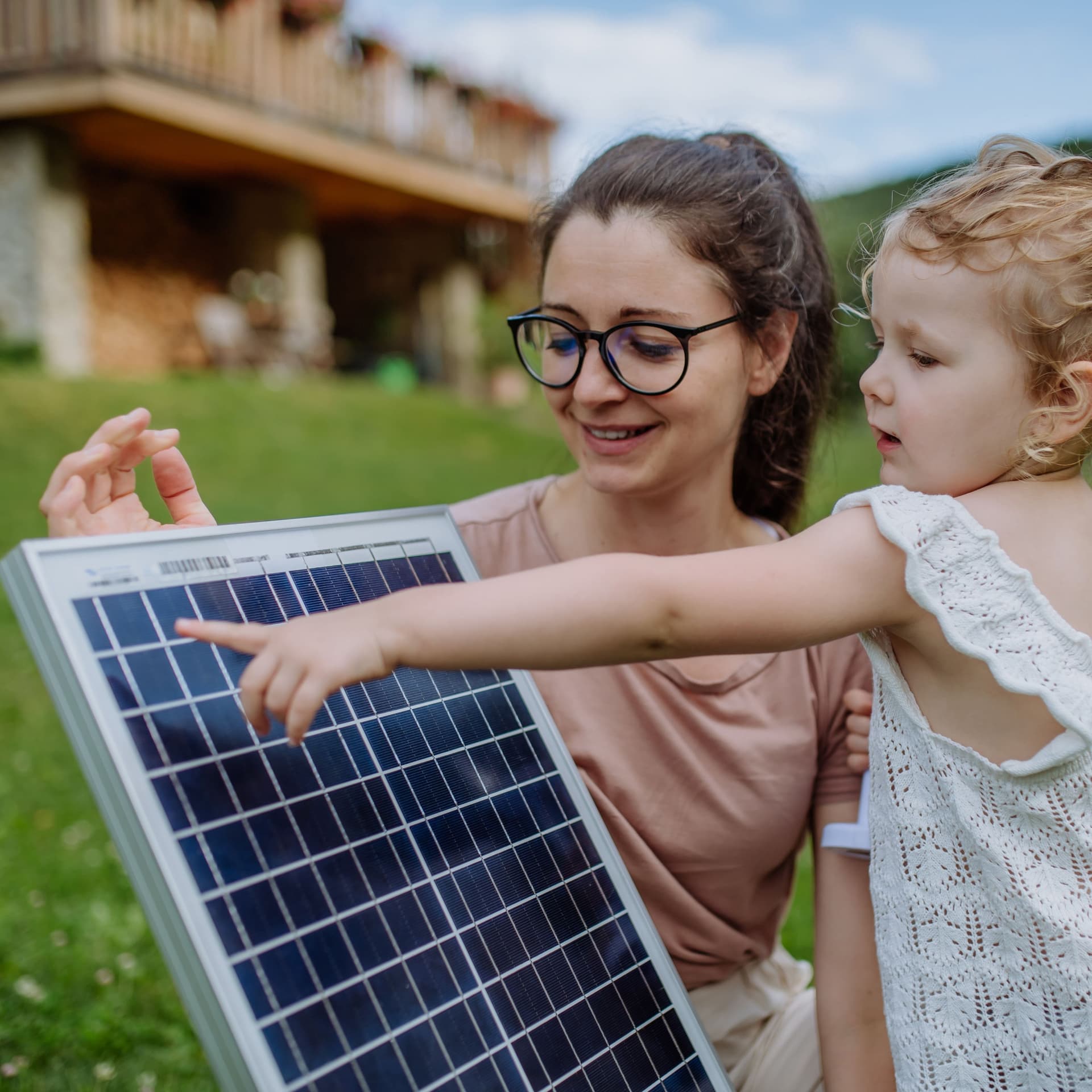 Eine junge Frau zeigt ihrer Tochter ein Solarpanel. Sie stehen auf einer Wiese. Im Hintergrund ist ein Haus zu sehen.