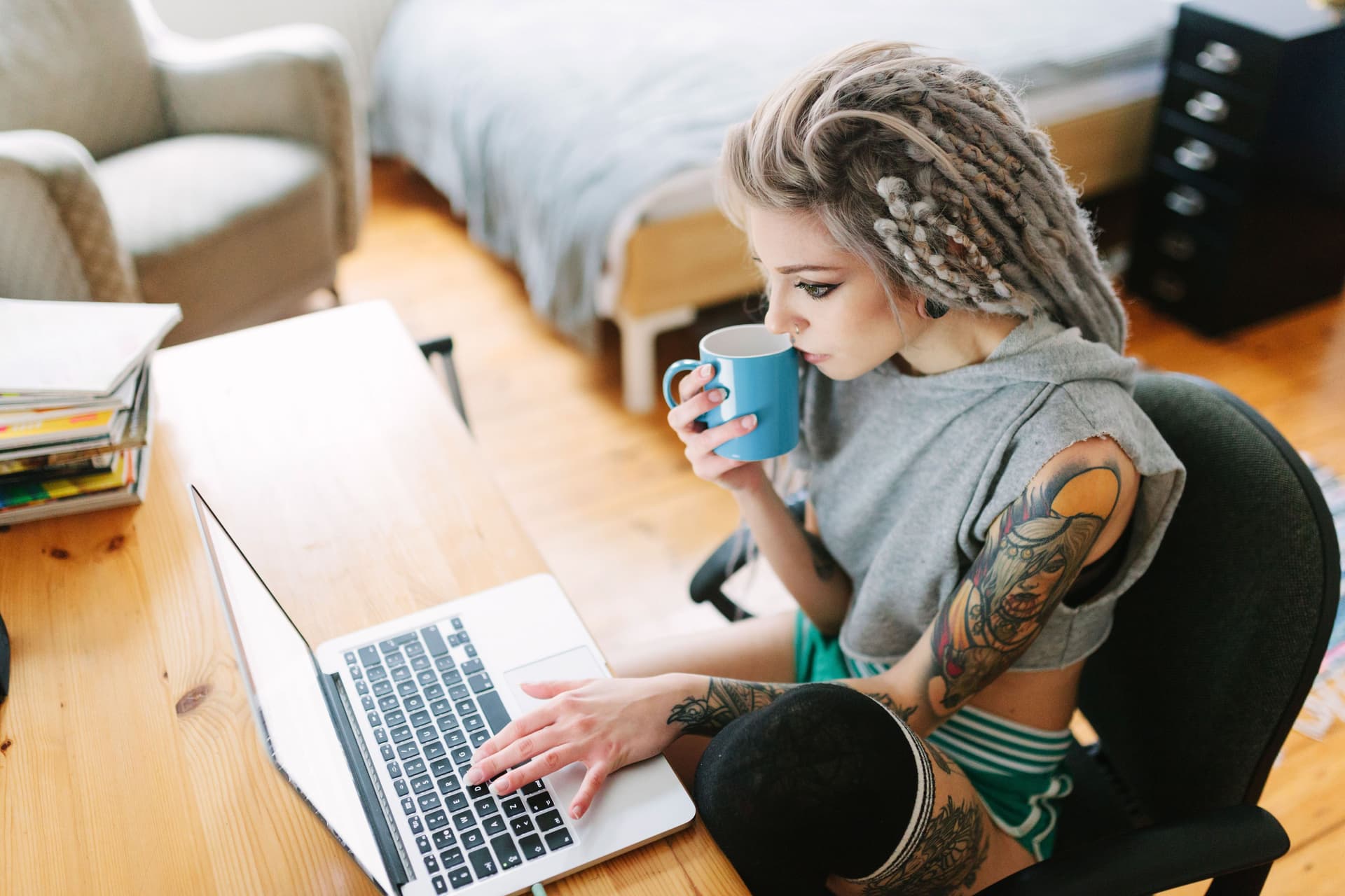 Eine junge tätowierte Frau mit grauen Dreadlocks sitzt konzentriert mit einer Tasse Kaffee vor ihrem Laptop.