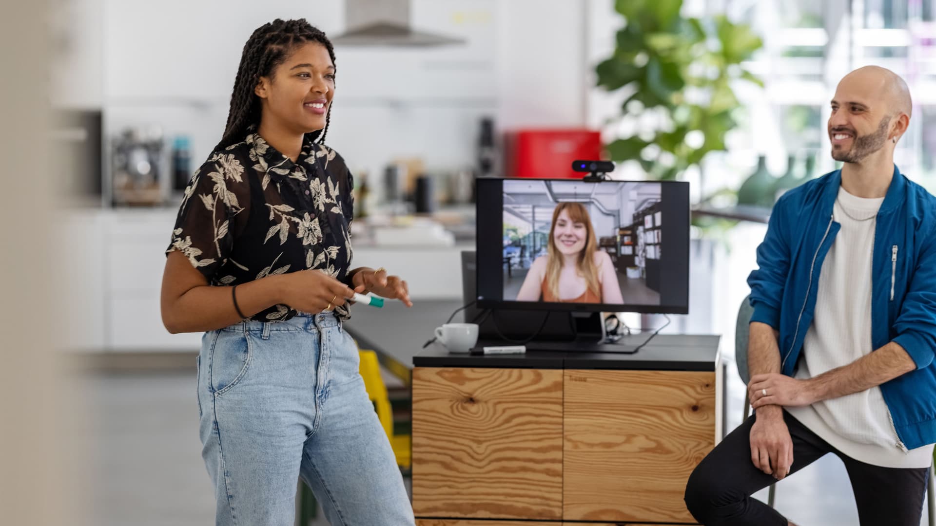 Eine POC und ein hellhäutiger Mann stehen in einer hybriden Arbeitssituation. Auf dem Bildschirm zwischen den beiden ist eine weiter Person zu sehen.