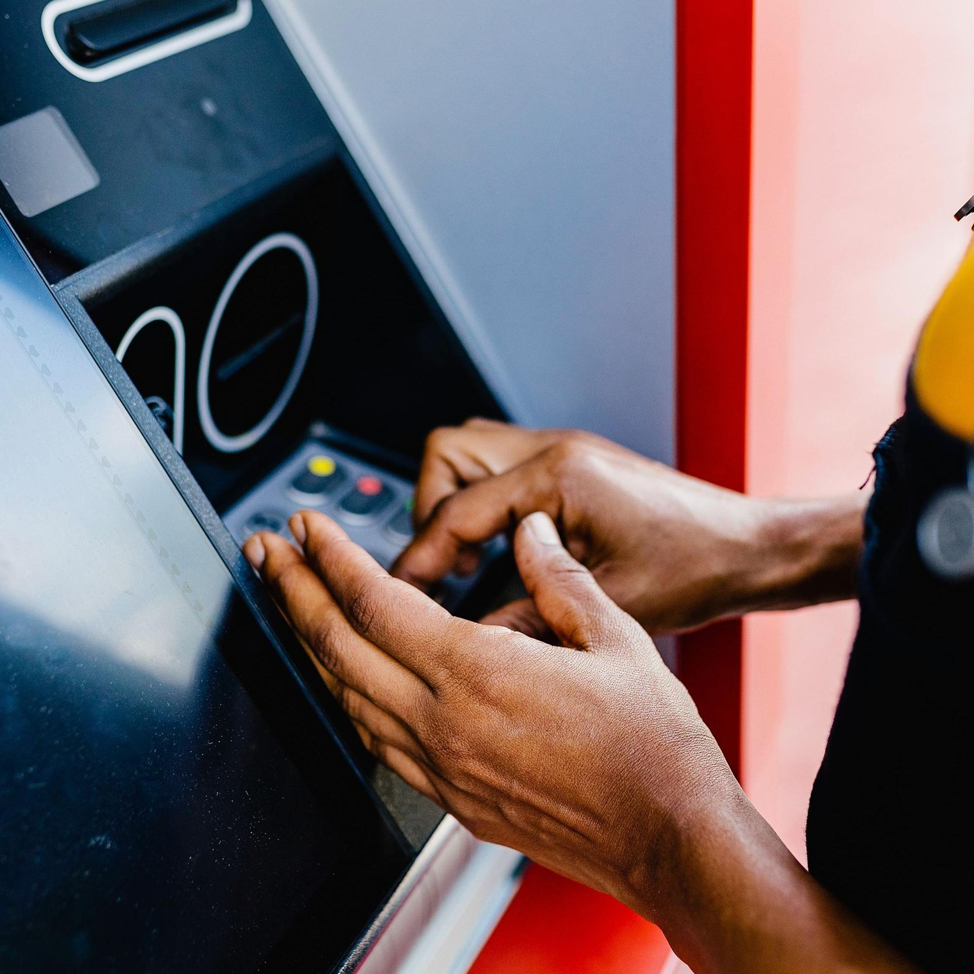 Nahaufnahme der Hände und Arme einer Frau, die mit verdeckter Hand ihren Pin-Code an einem Bankautomaten eingibt.