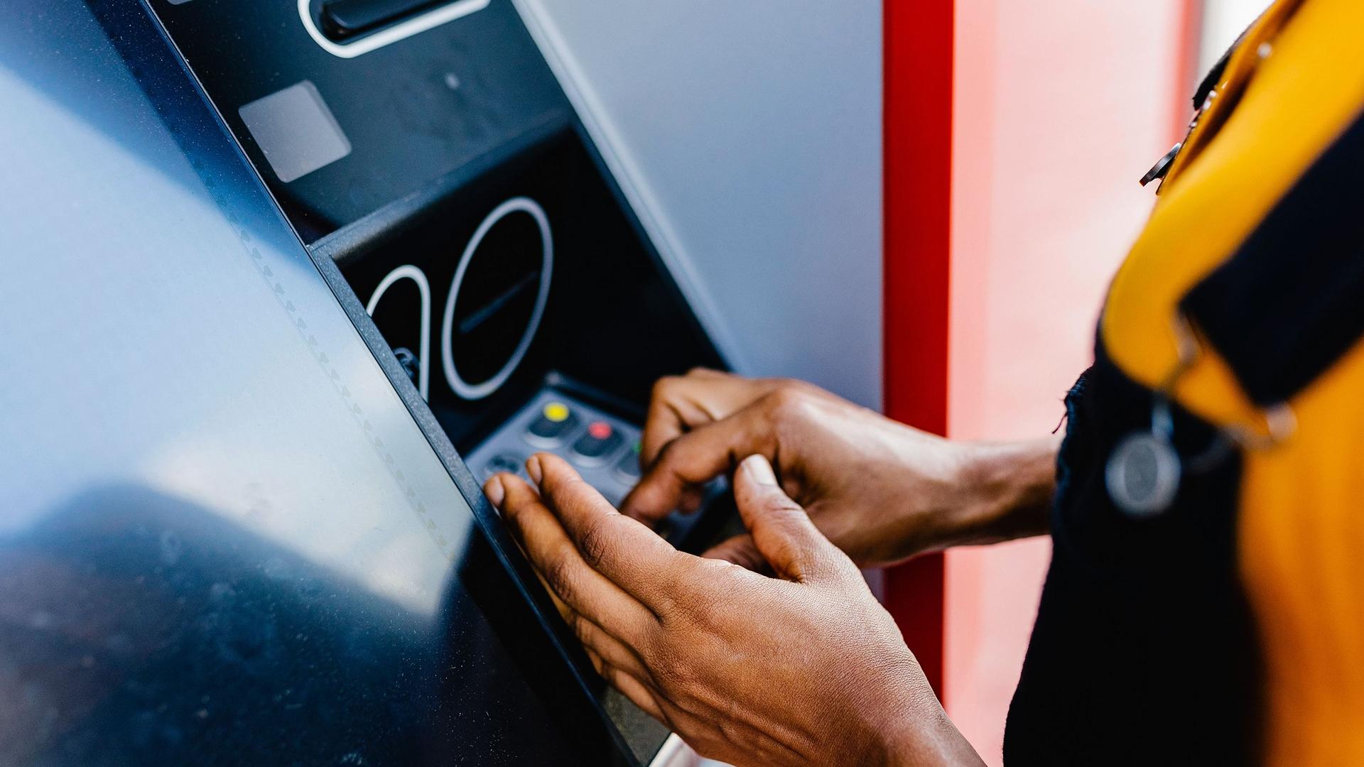 Nahaufnahme der Hände und Arme einer Frau, die mit verdeckter Hand ihren Pin-Code an einem Bankautomaten eingibt.