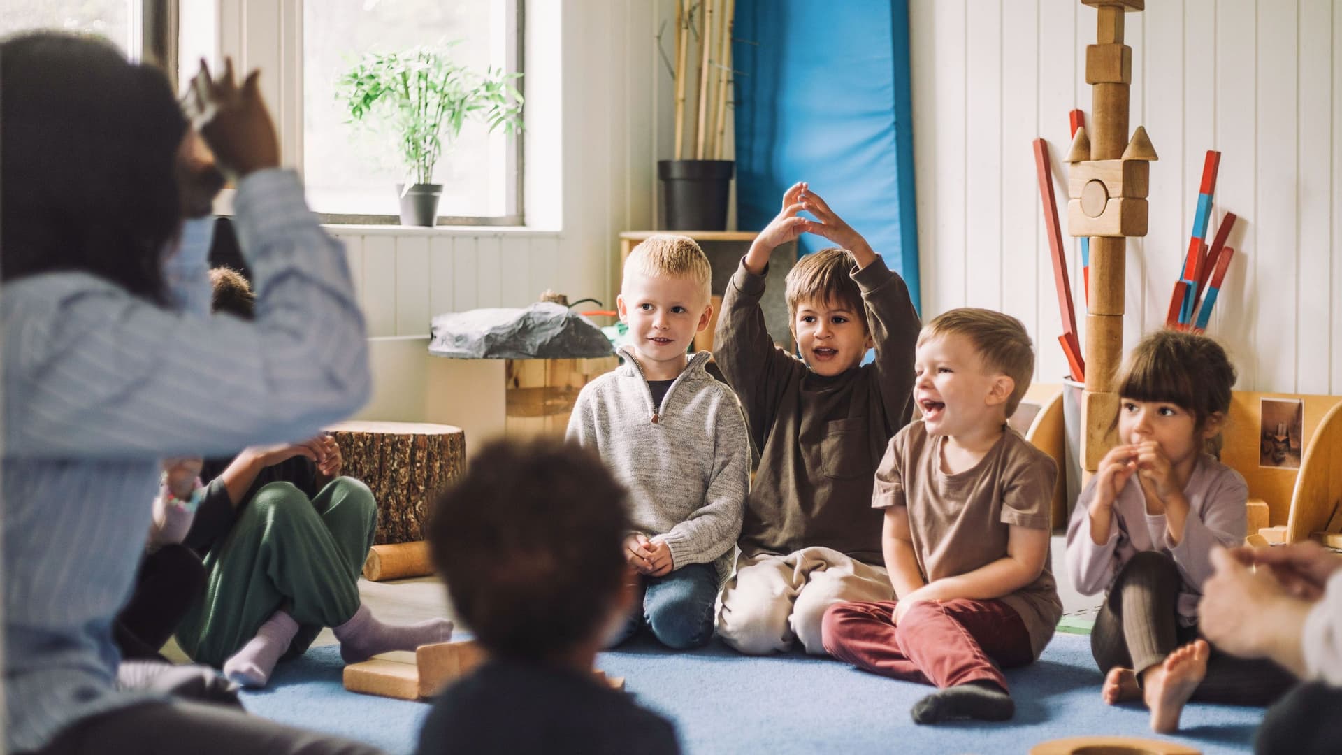 Eine Erzieherin sitzt in einer Kindertagesstaette mit einer Kindergruppe in einem Sitzkreis.