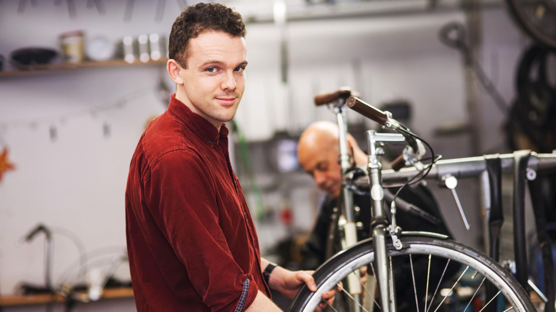Ein junger Fahrradtechniker, der in einer Werkstatt an dem Vorderrad eines Fahrrads arbeitet und in die Kamera lächelt.