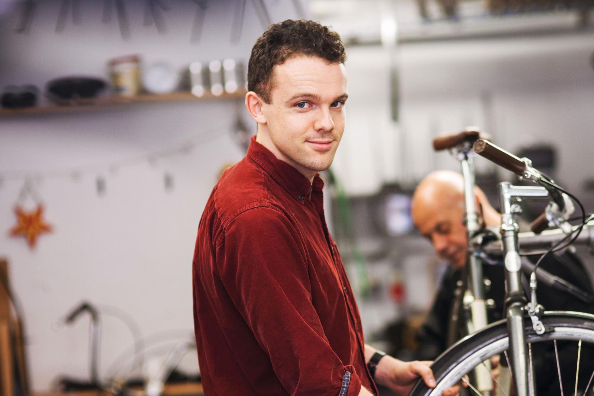 Ein junger Fahrradtechniker, der in einer Werkstatt an dem Vorderrad eines Fahrrads arbeitet und in die Kamera lächelt.