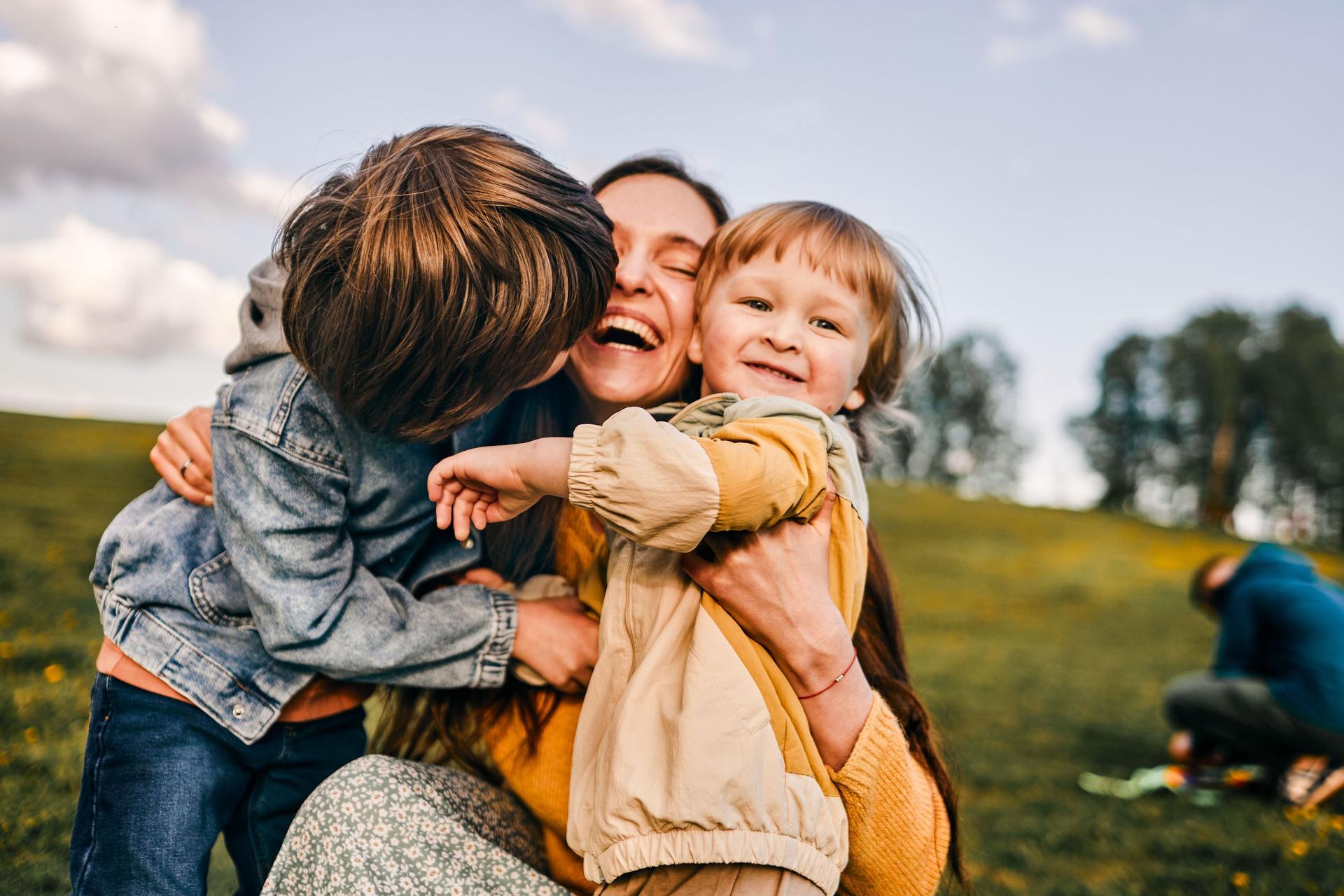 Eine junge Mutter umarmt lachend ihre zwei Kinder auf einer Wiese.