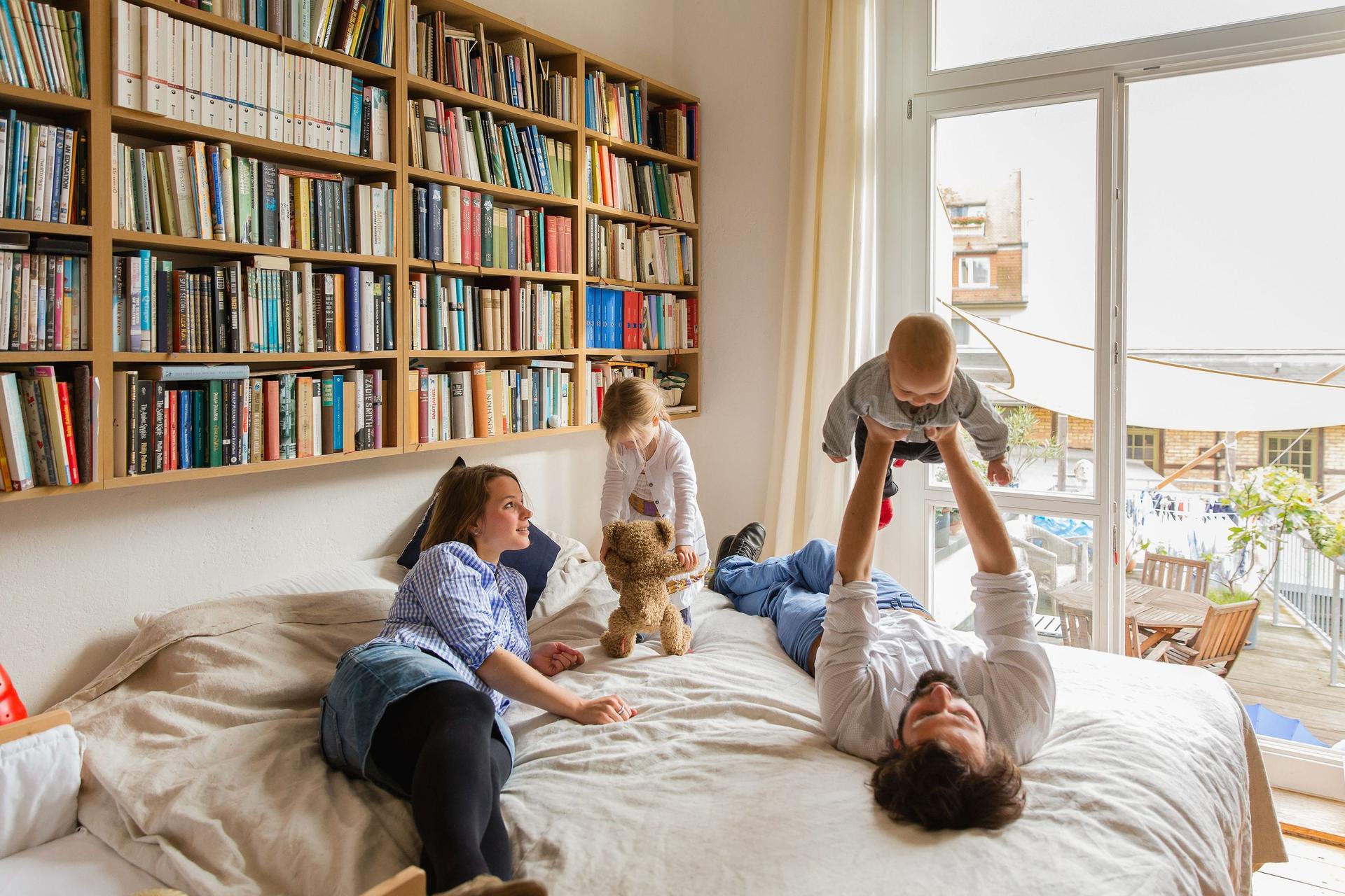 Eine vierköpfige Familie spielt zusammen auf einem Bett. Über dem Bett hängt ein großes Bücherregal.