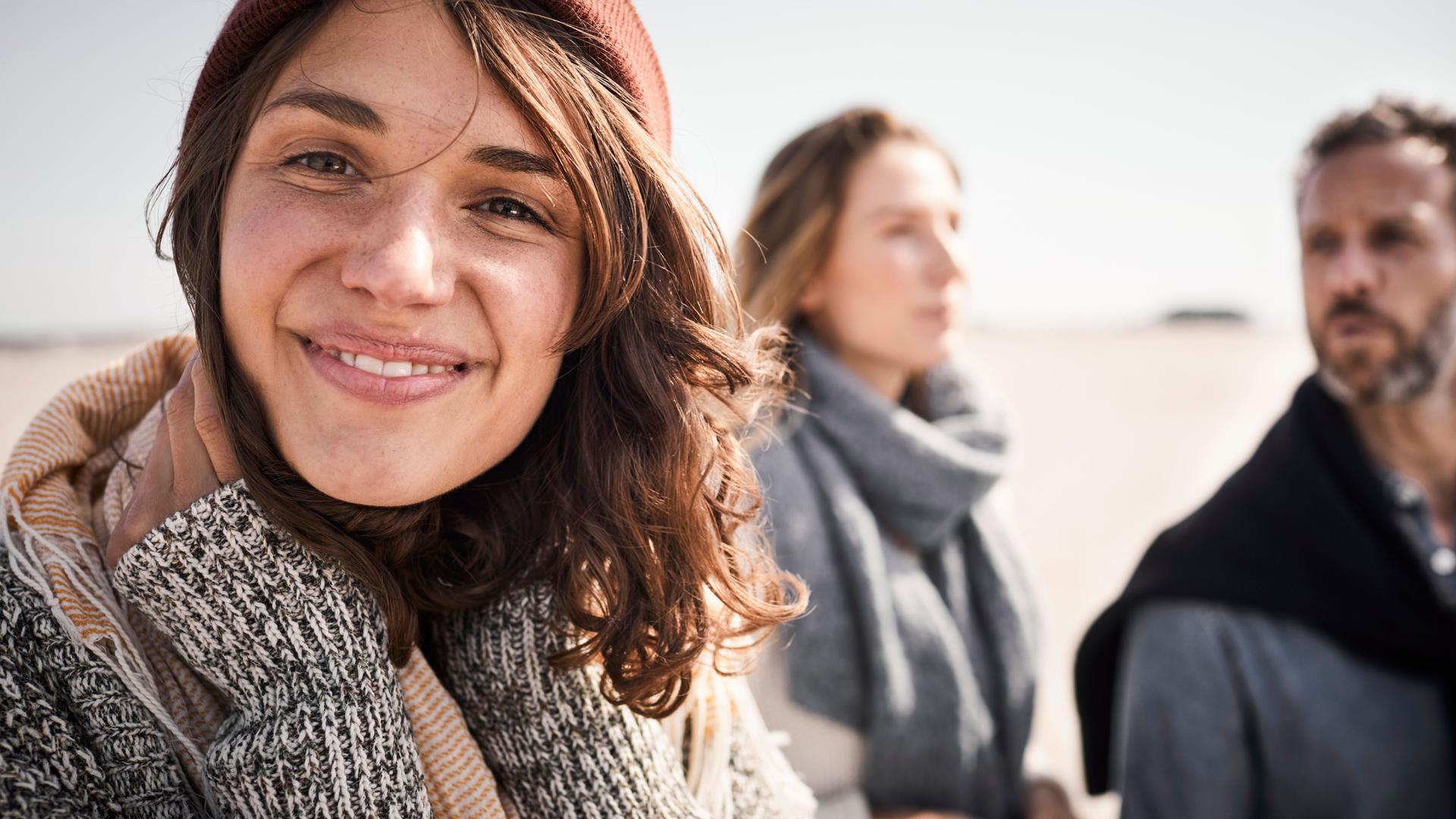 Porträt einer jungen Frau am Strand, die in die Kamera lächelt. Hinter ihr stehen eine Frau und ein Mann.