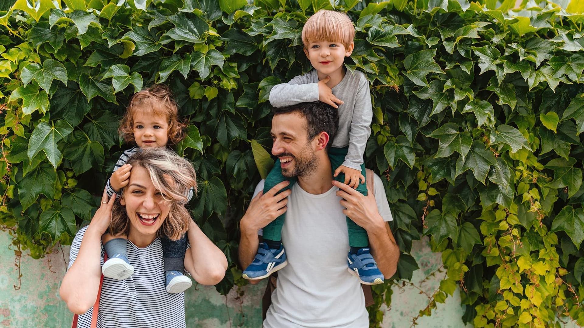 Eine sommerlich gekleidete Mutter und ein Vater haben jeweils ein Kind auf ihren Schultern. Sie stehen vor einer begrünten Wand und lächeln.