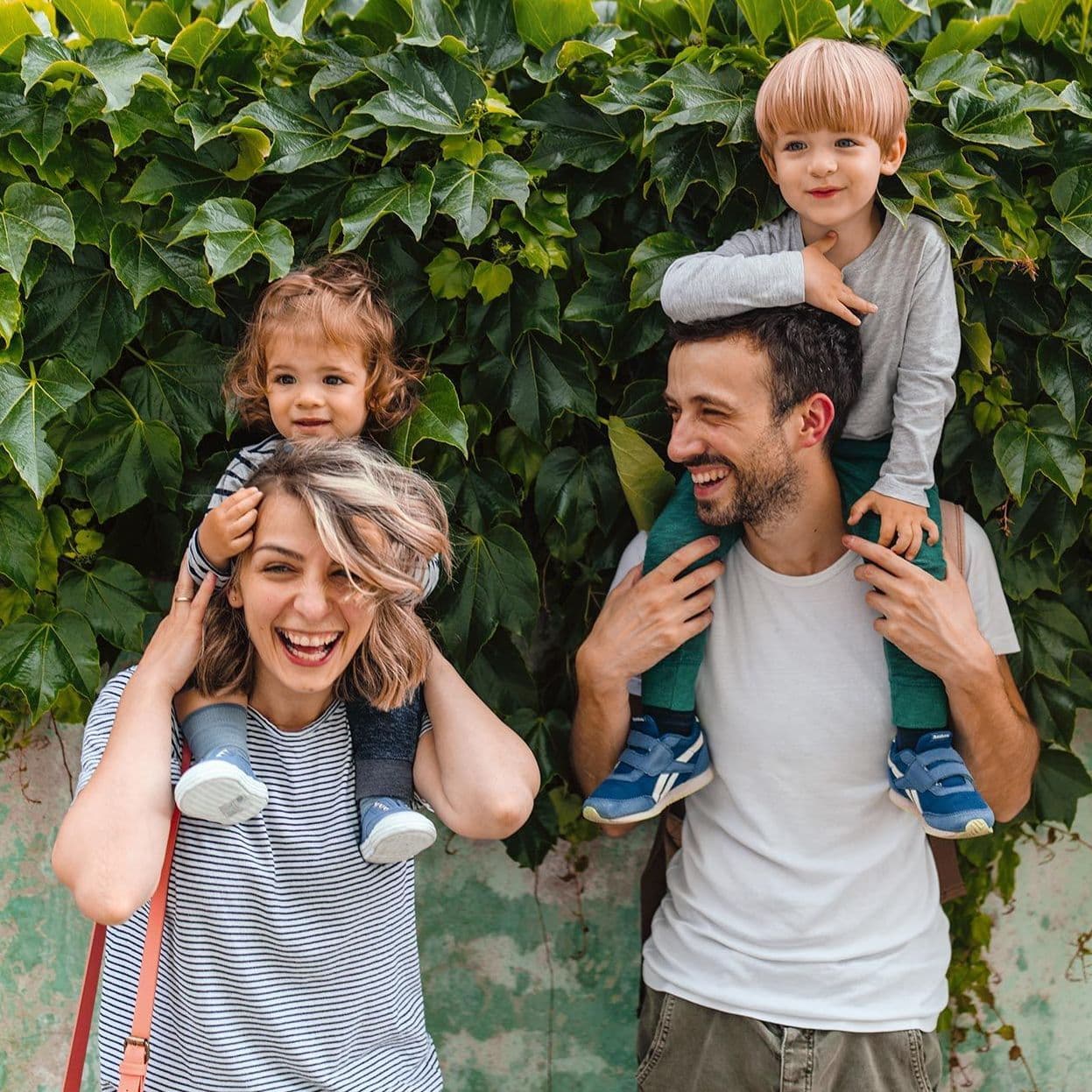 Eine sommerlich gekleidete Mutter und ein Vater haben jeweils ein Kind auf ihren Schultern. Sie stehen vor einer begrünten Wand und lächeln.