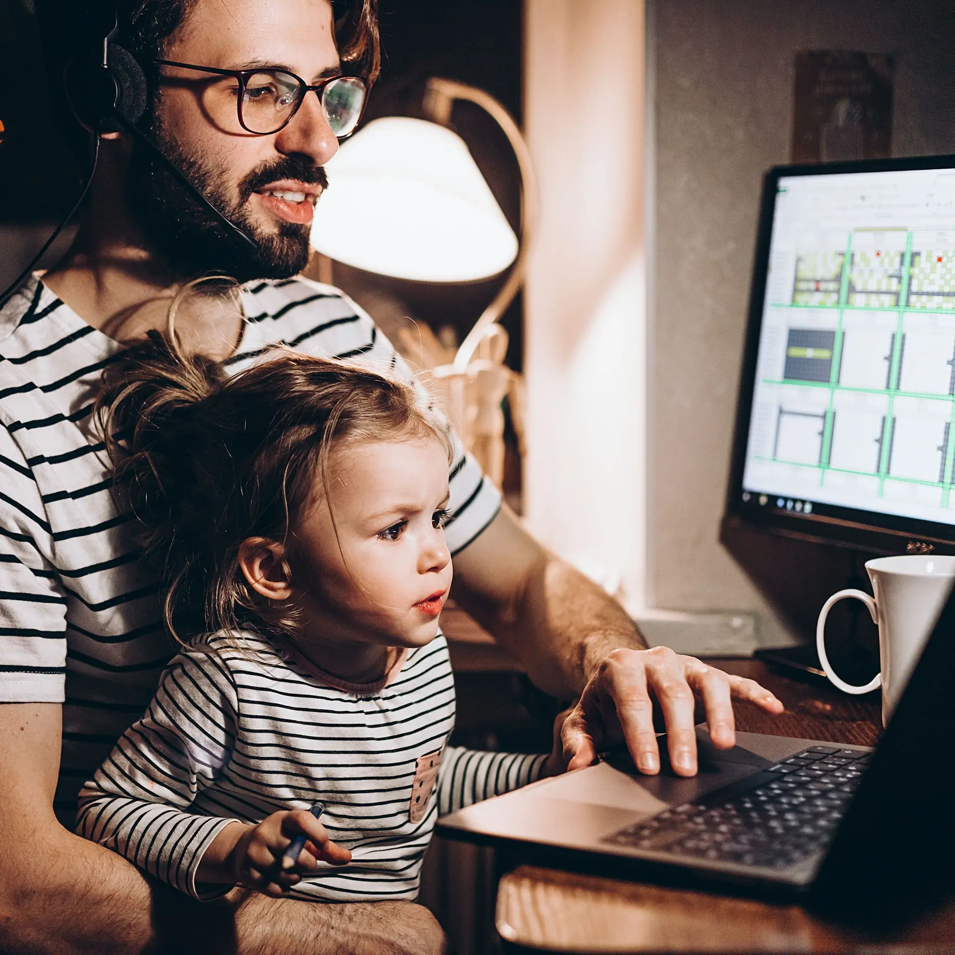 Ein mittelalter Vater sitzt mit einem kleinen Mädchen auf dem Schoß vor einem Laptop und Monitor im Homeoffice.