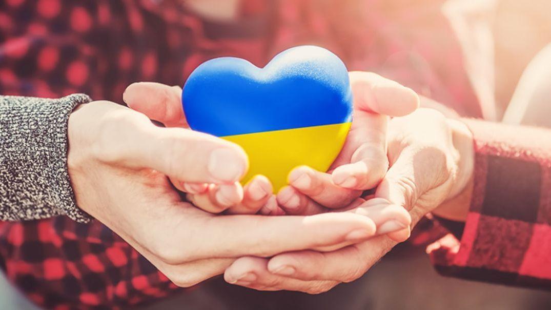 Blau-gelbes Herz aus Stein in zwei ineinander gefalteten Händen, die Farben symbolisieren die Flagge der Ukraine