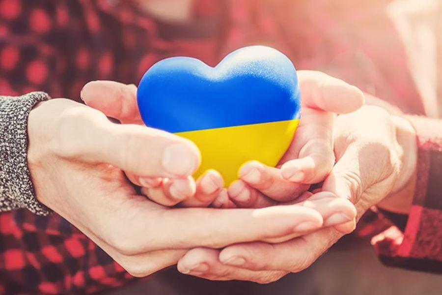 Blau-gelbes Herz aus Stein in zwei ineinander gefalteten Händen, die Farben symbolisieren die Flagge der Ukraine