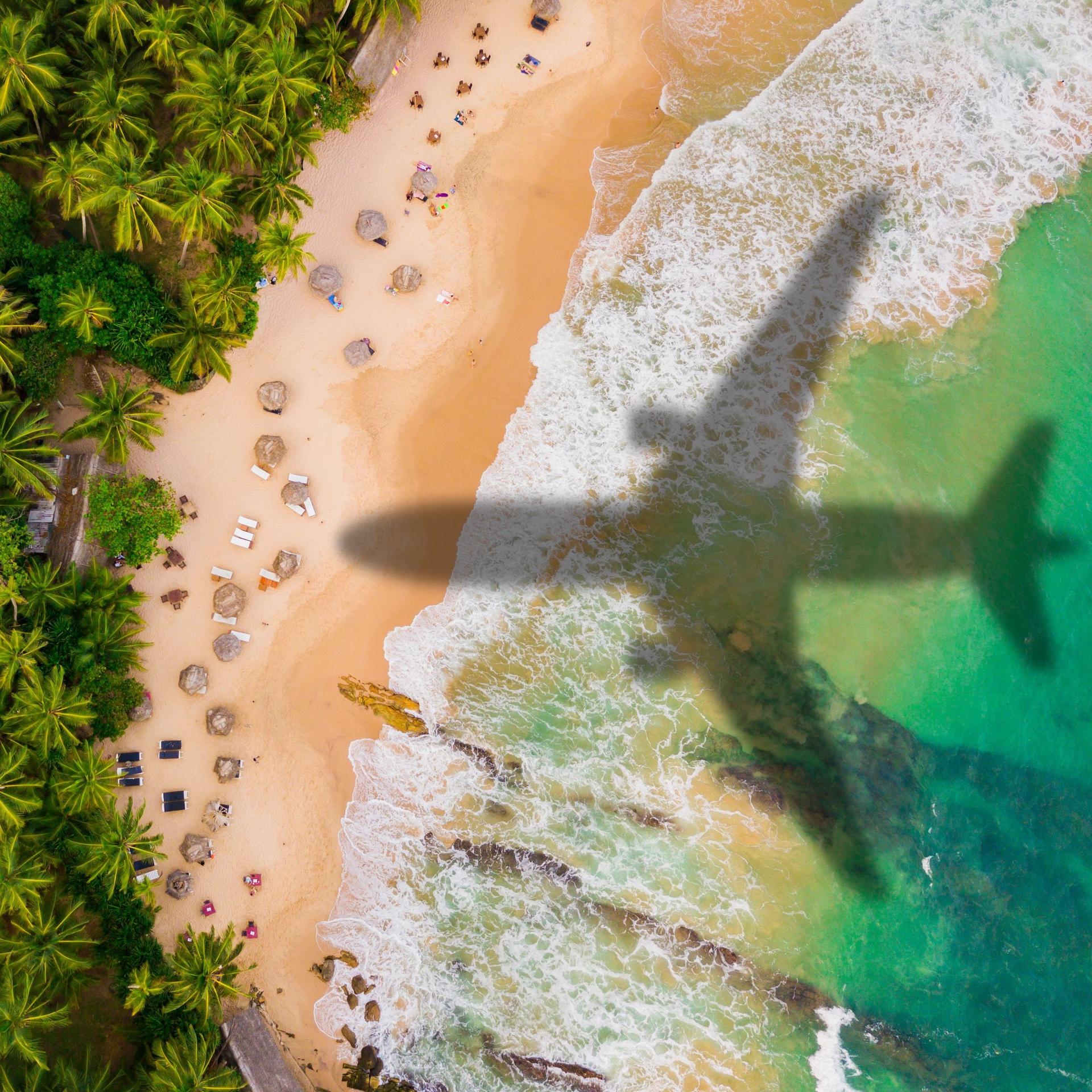 Ein Flugzeug fliegt über einen karibischen Strand. Man sieht nur den Schatten des Flugzeugs auf dem Strand.
