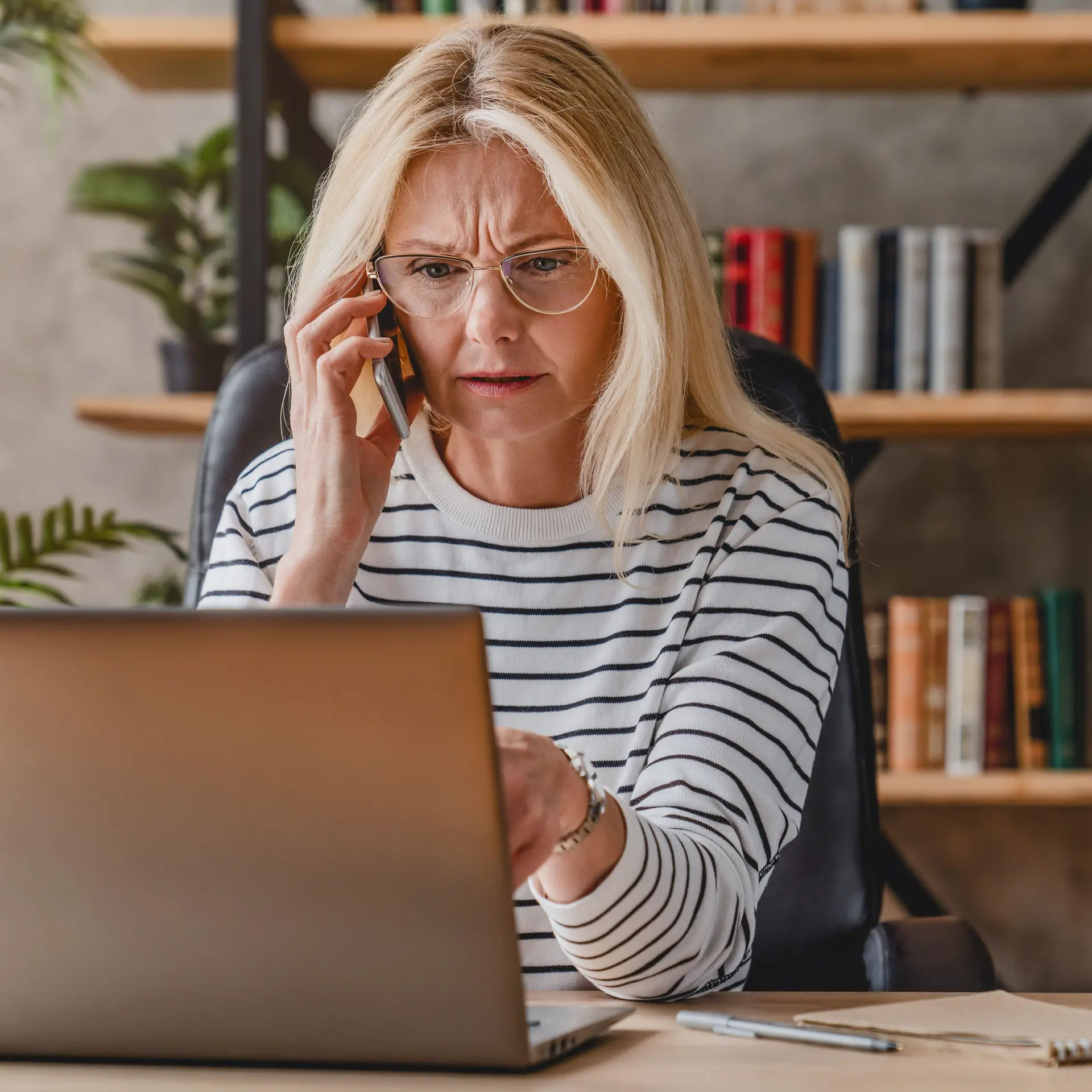 Reife Frau mit Streifenpulli sieht gestresst aus, während sie einen Laptop benutzt und mit einem Smartphone telefoniert.