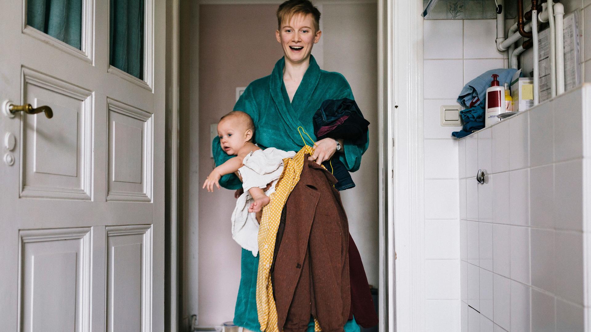 Eine junge Frau, die einige Kleider und ein Baby in ein Badezimmer trägt. Sie trägt einen Bademantel und lächelt mit geöffnetem Mund.
