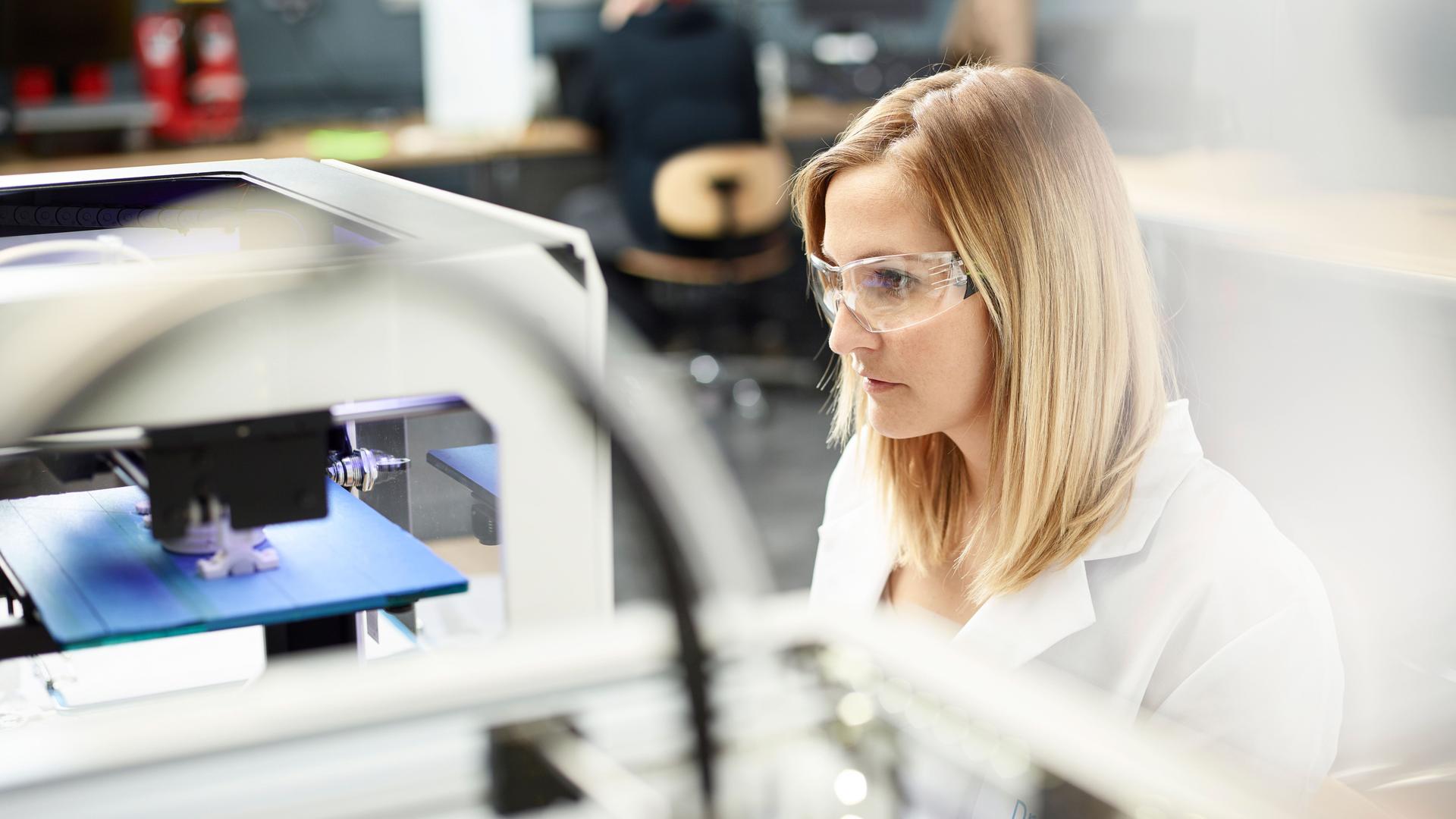 Junge Frau mit Schutzbrille und weißem Kittel sitzt vor einem 3D Printer und bedient diesen konzentriert
