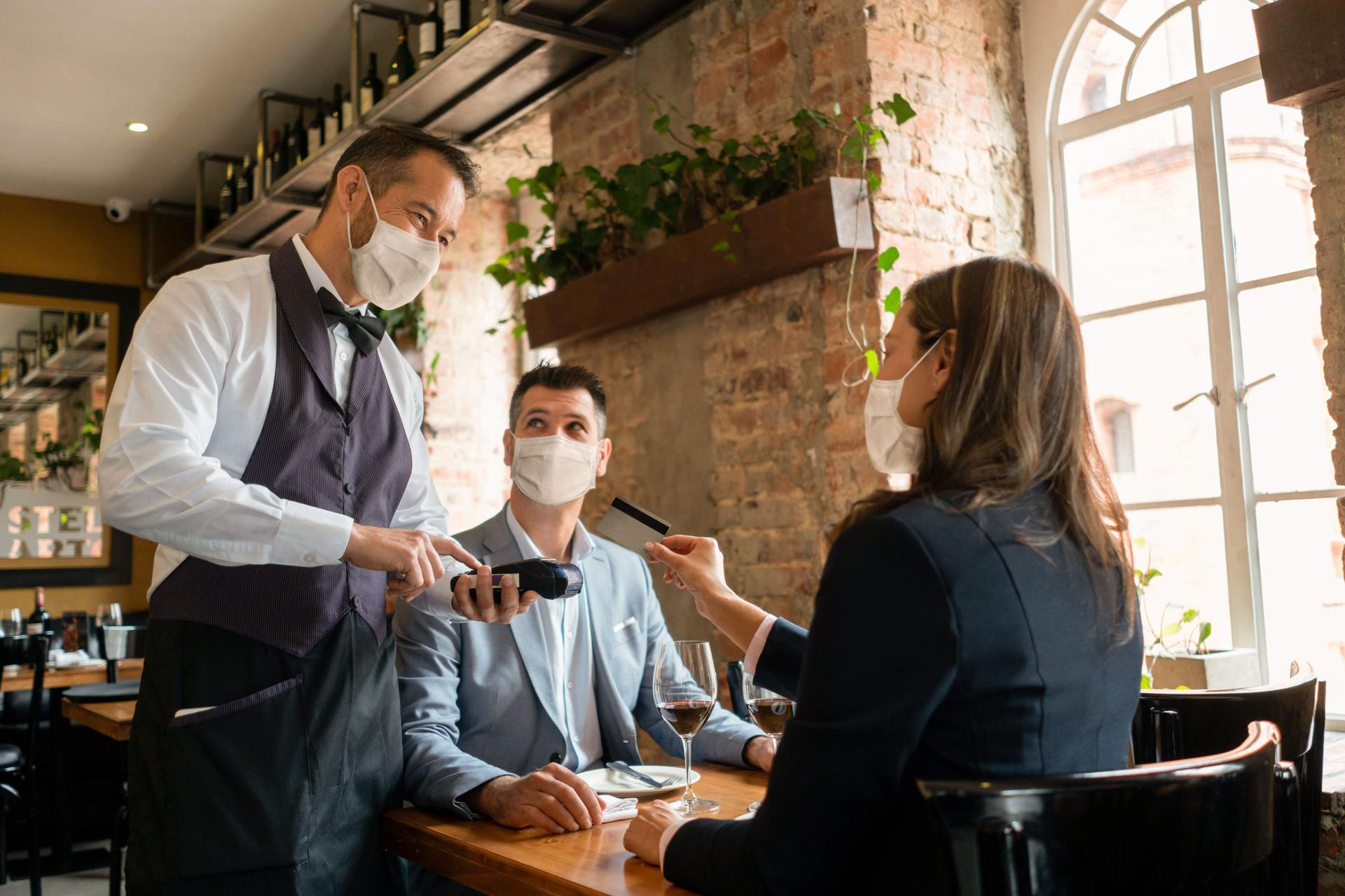 Eine Frau und ein Mann sitzen in einem Restaurant an einem Tisch. Die Frau reicht dem Kellner, der am Tisch steht, eine Bankkarte. Alle tragen eine Mund-Nasen Bedeckung.