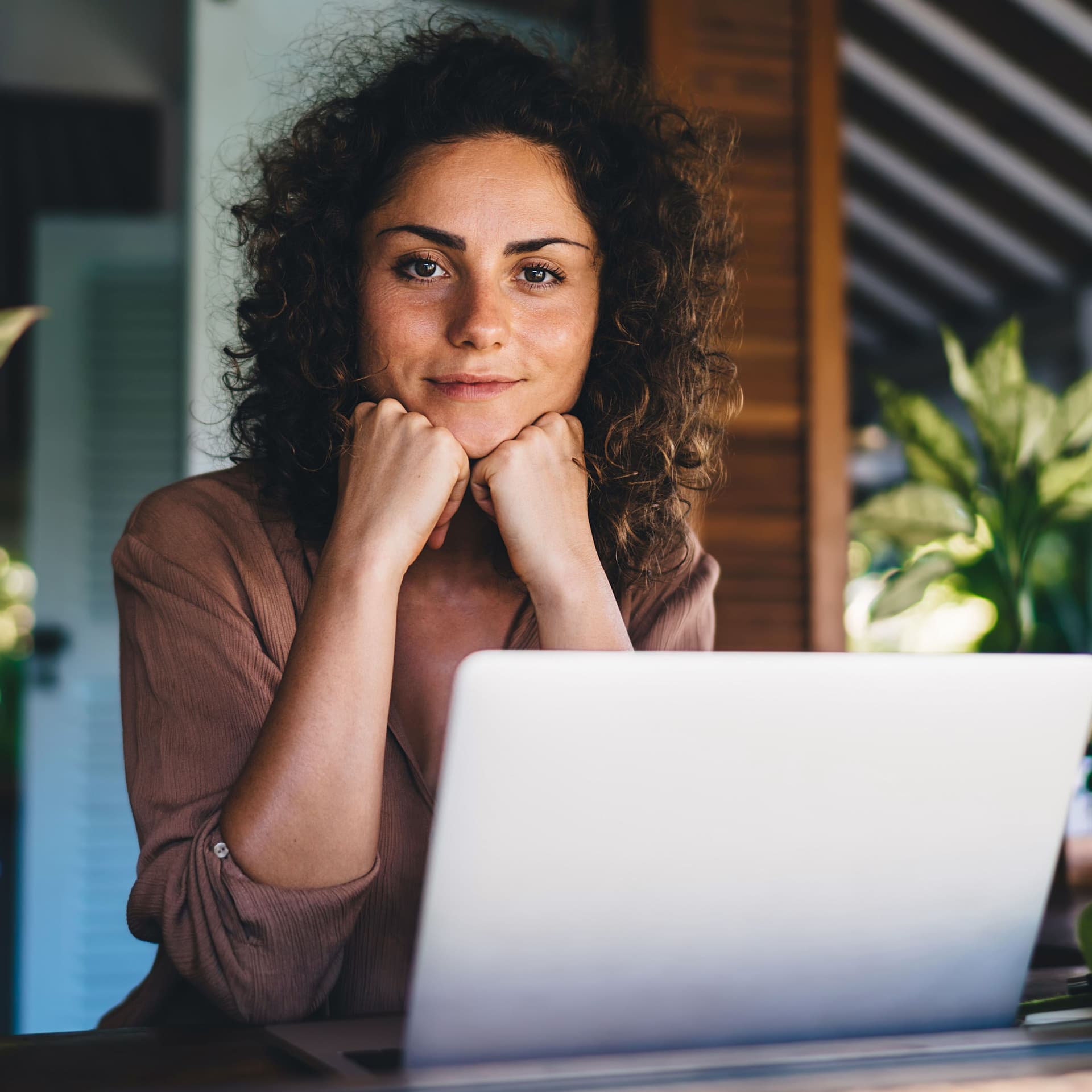 Junge Frau sitzt auf einer Terrasse mit Pflanzen vor einem Laptop. Sie stützt ihren Kopf auf die Hände und lächelt in die Kamera.