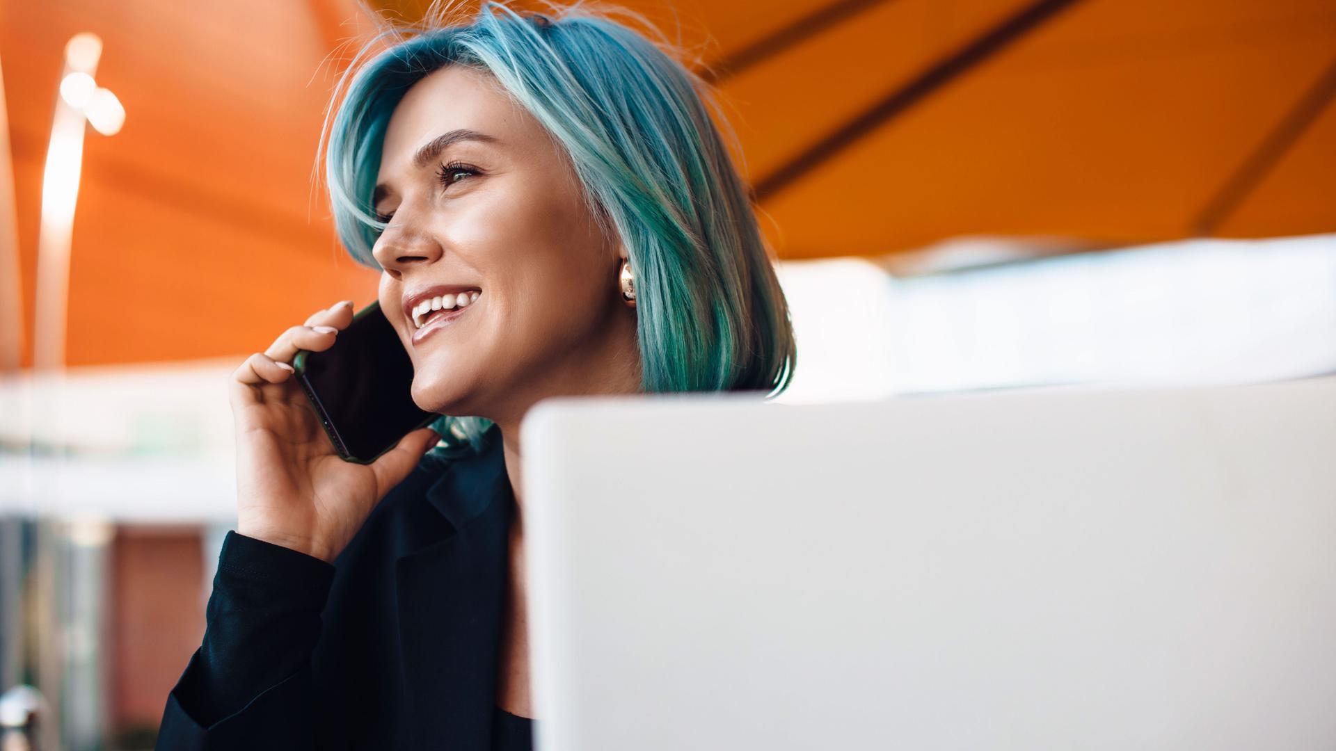 Junge lächelnde Frau mit blauen Haaren am Telefonieren mit Laptop vor sich.