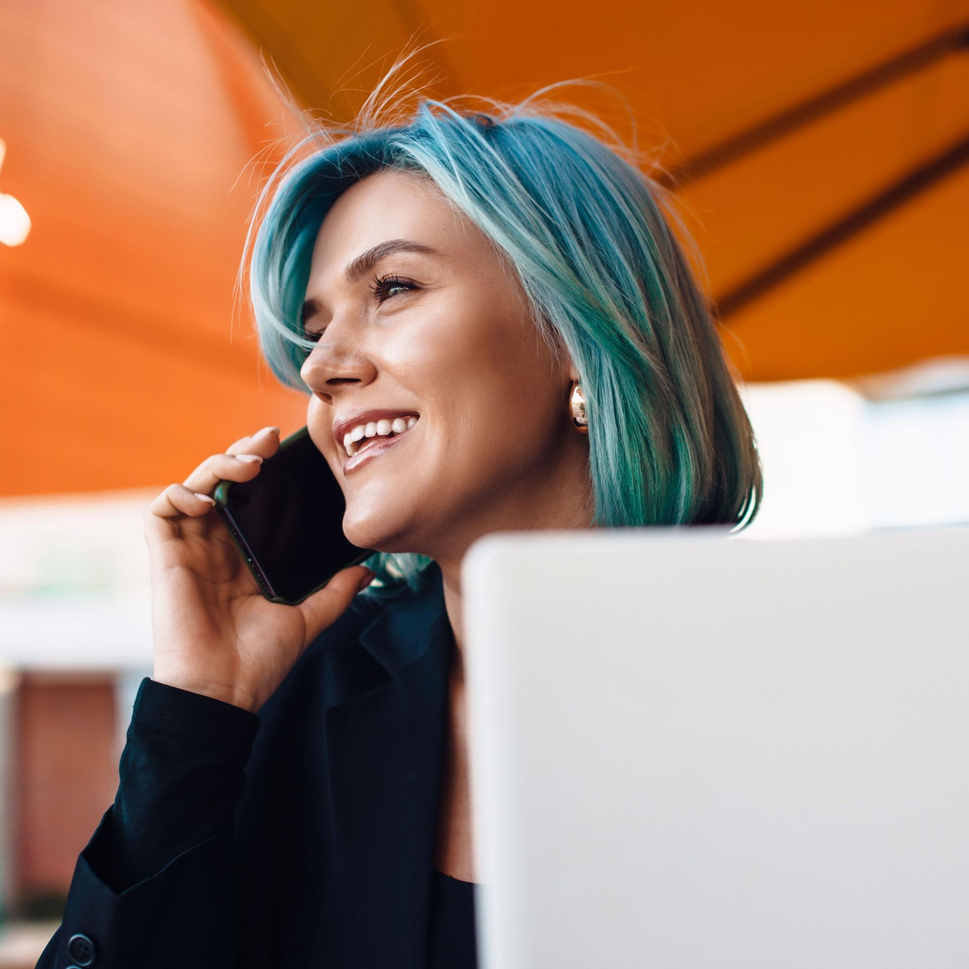 Junge lächelnde Frau mit blauen Haaren am Telefonieren mit Laptop vor sich.