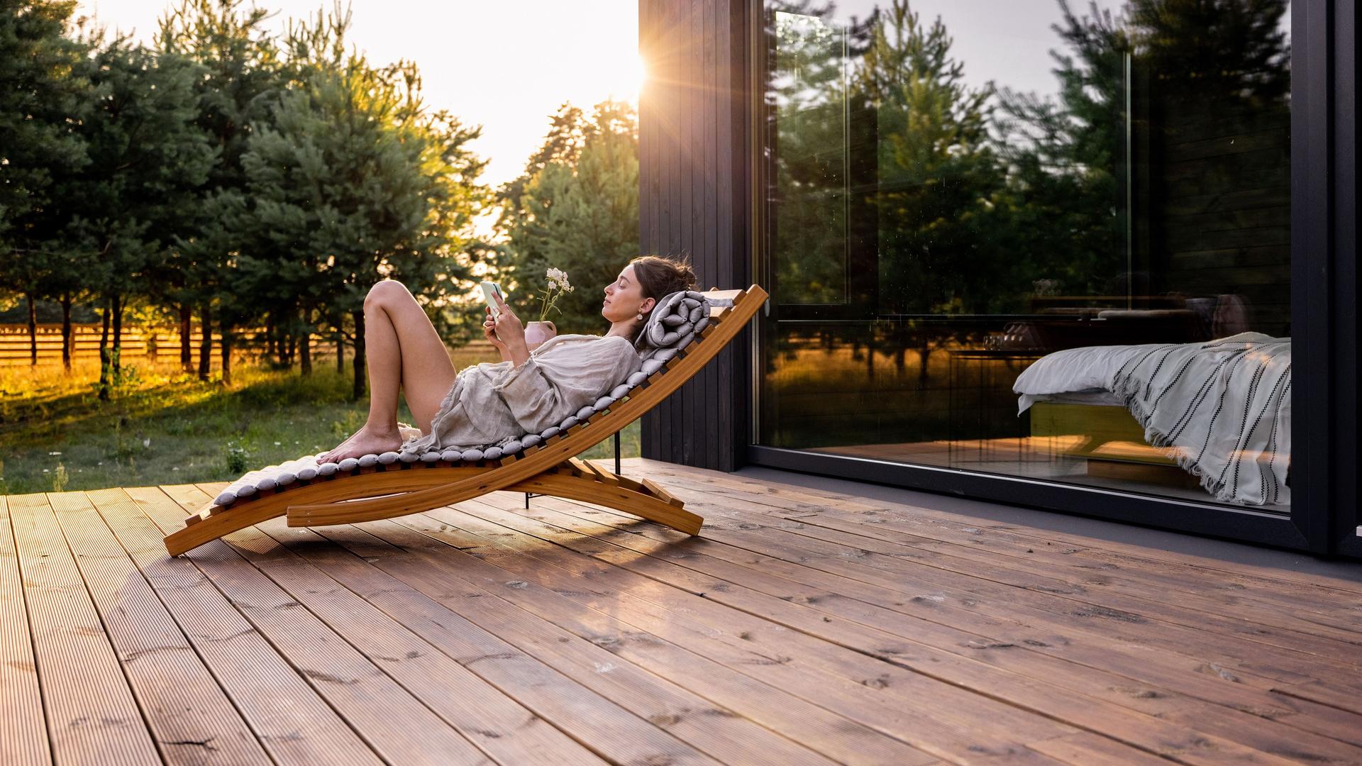 Frau liegt auf einer Sonnenliege auf der Holzterrasse eines Hauses. 