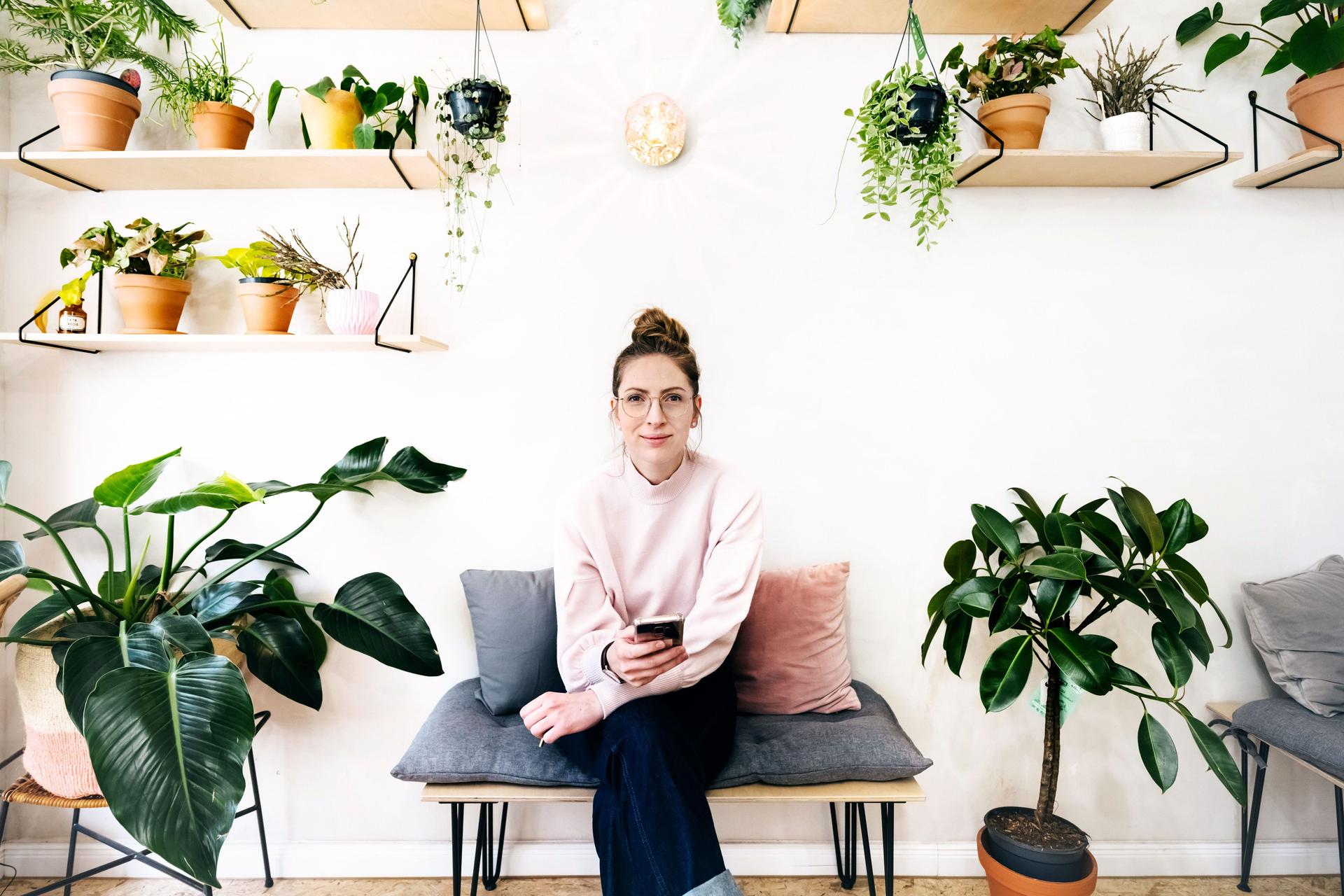 Eine junge Frau mit Smartphone in der Hand sitzt auf einer Bank mit grauem Kissen in einem hellen Raum. Sie guckt in die Kamera und ist umgeben von vielen Pflanzen an der Wand und am Boden.