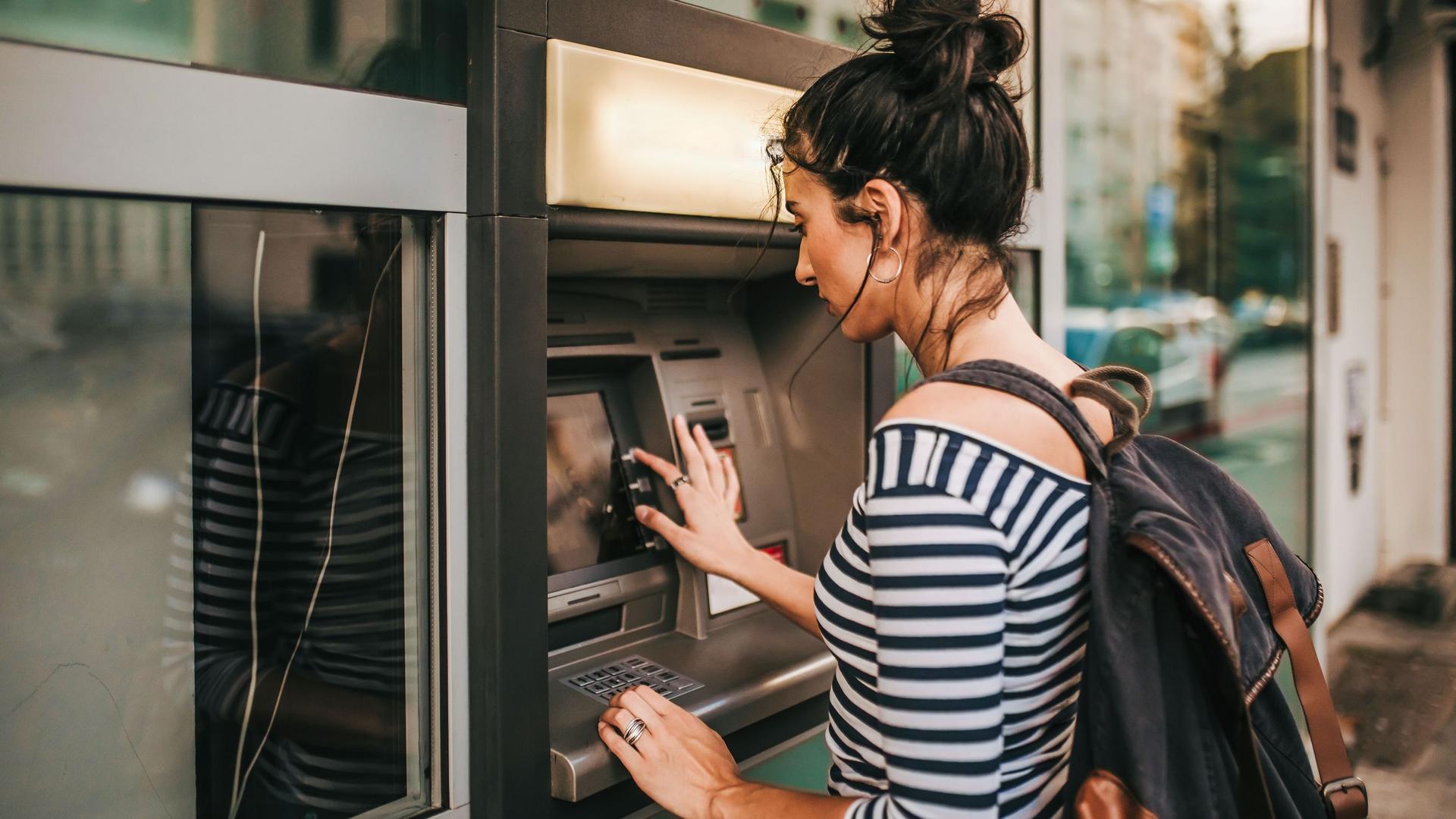 Eine junge Frau mit Rucksack und einem geringelten Shirt hebt Geld von einem Bankautomaten auf der Straße ab.