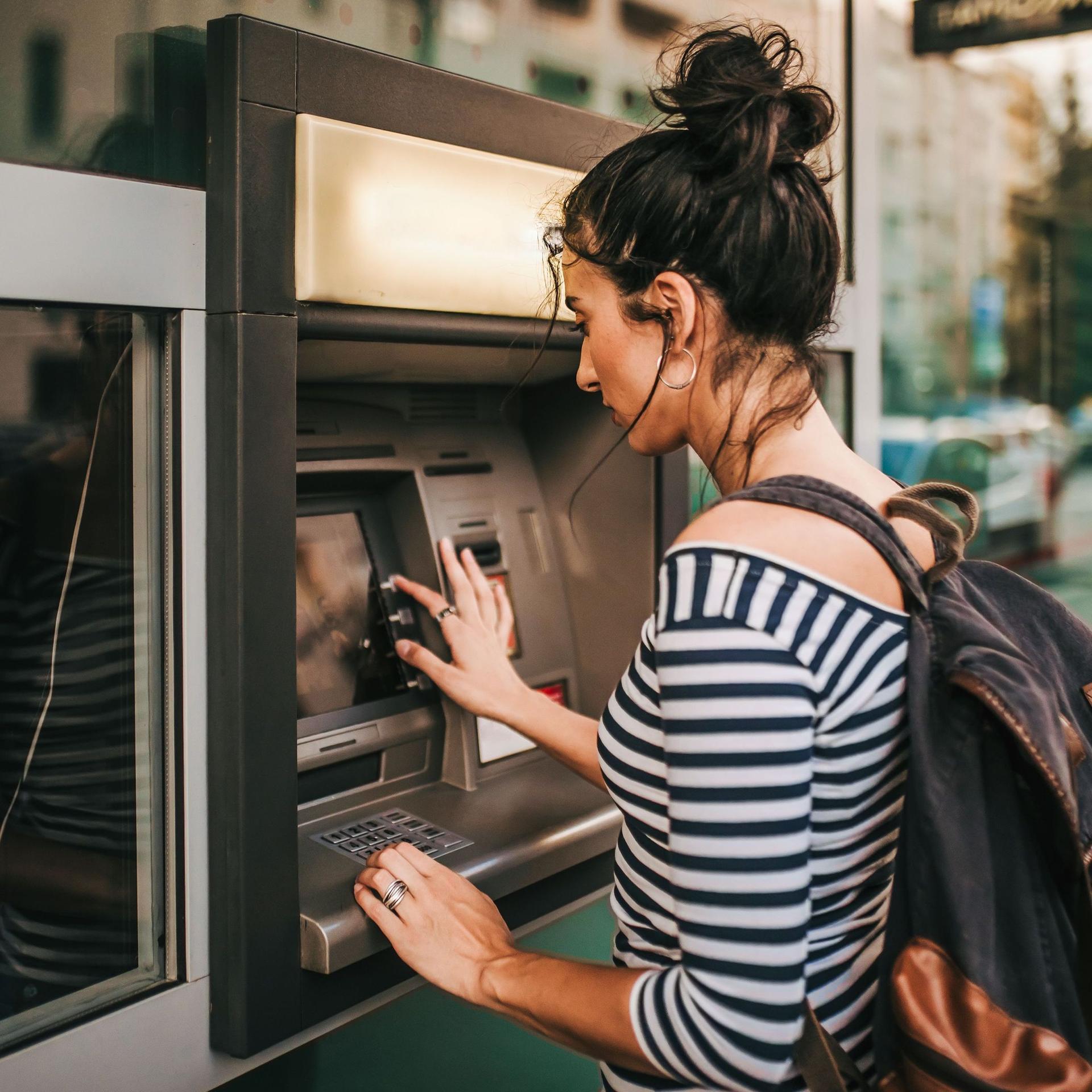 Eine junge Frau mit Rucksack und einem geringelten Shirt hebt Geld von einem Bankautomaten auf der Straße ab.