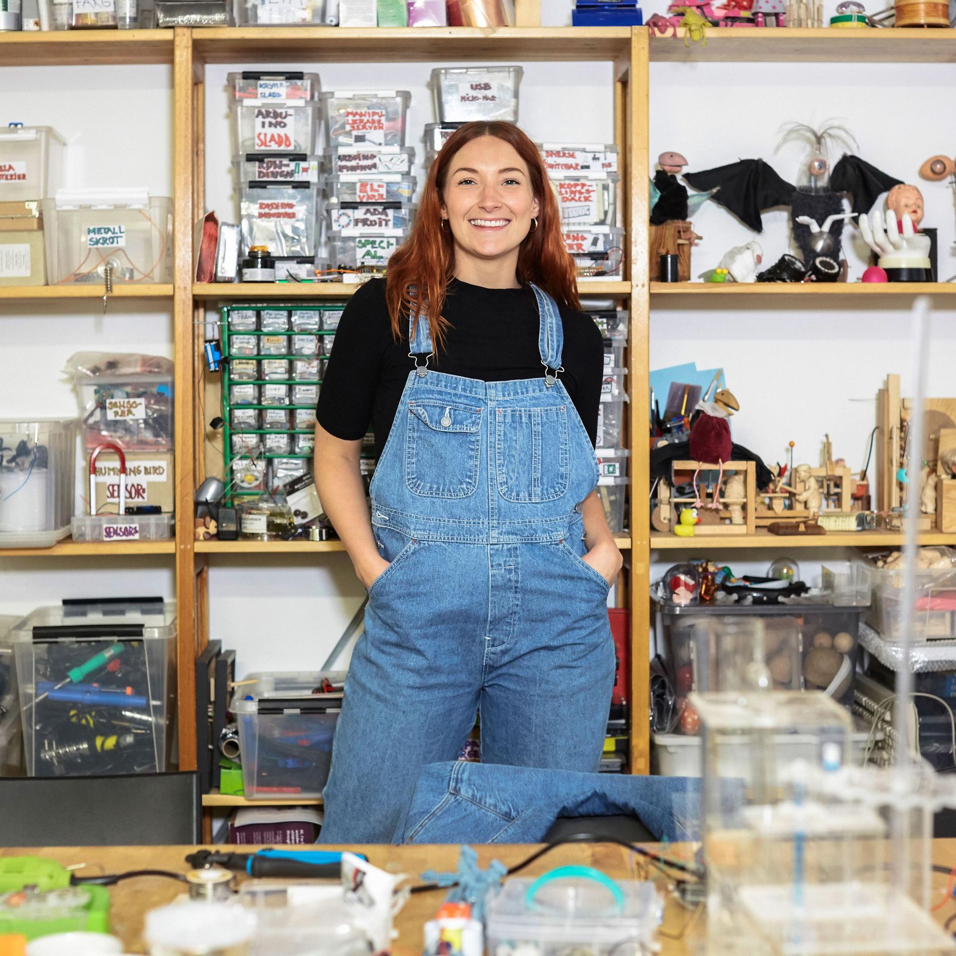 Porträt einer jungen Frau, die mit den Händen in den Taschen vor dem Regal in ihrer Werkstatt steht.