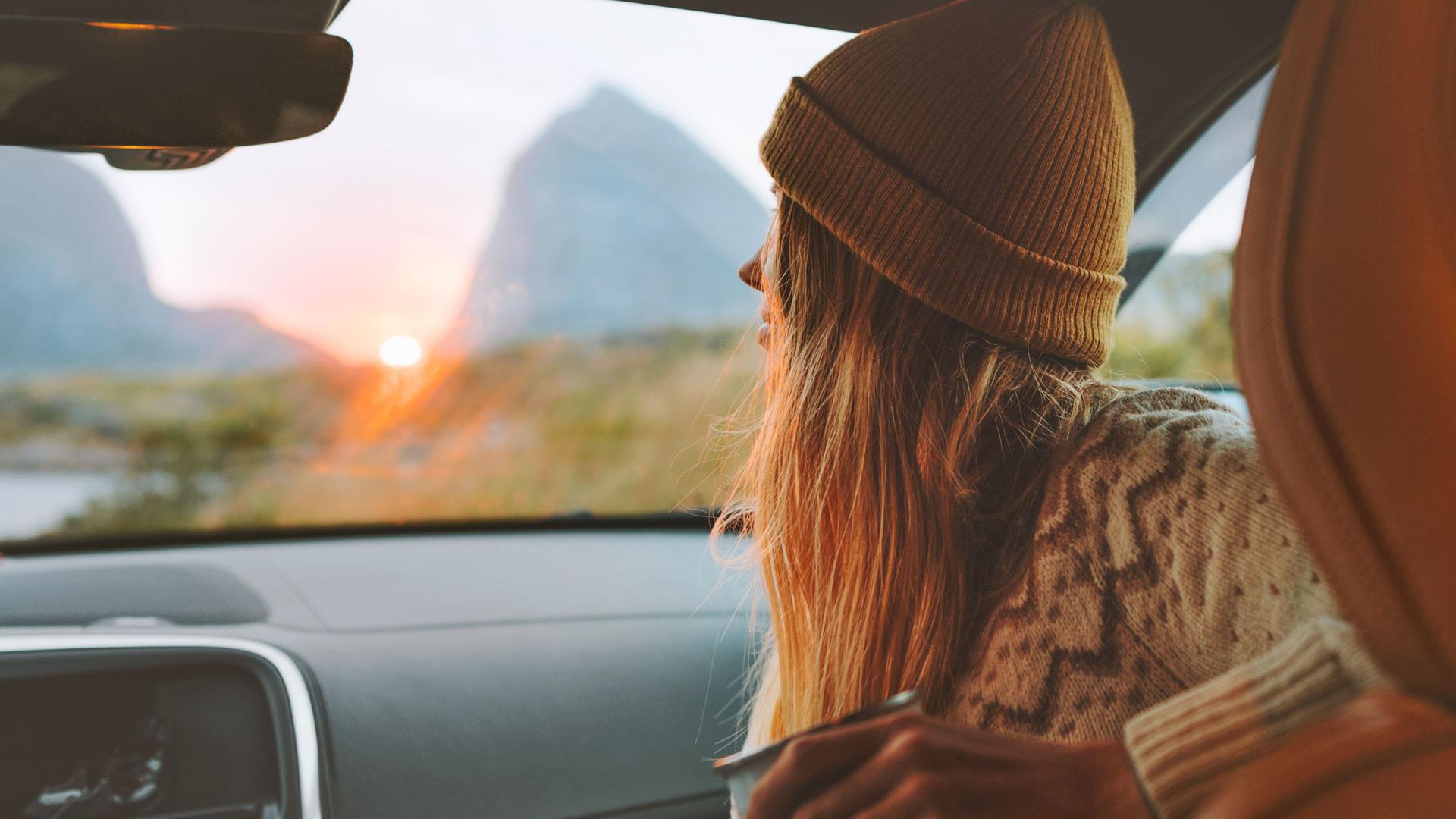 Junge Frau sitzt im Auto und durch die Frontscheibe. Im Hintergrund sind Berge und ein Sonnenuntergang zu sehen.