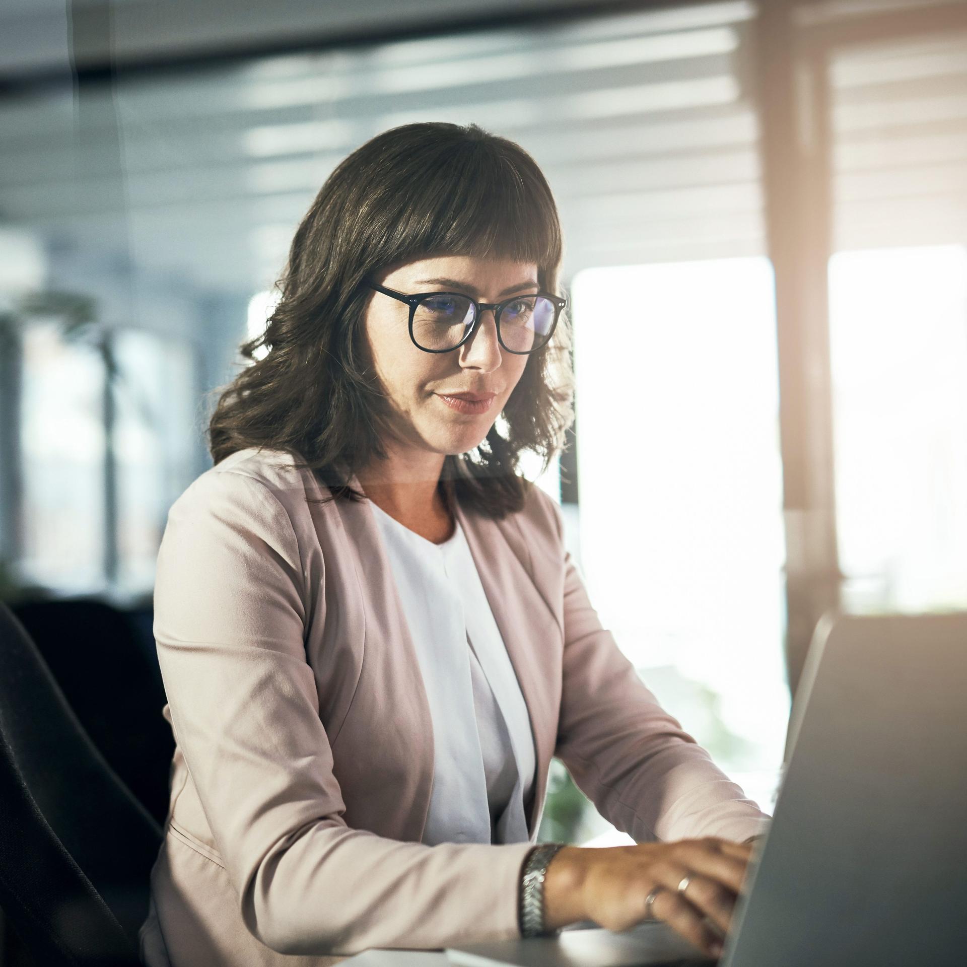Seitenansicht einer Frau mit Brille mittleren Alters, die in einer abendlichen Lichtstimmung alleine vor einem Laptop sitzt. Im Hintergrund ist ein modernes Büro erkennbar.