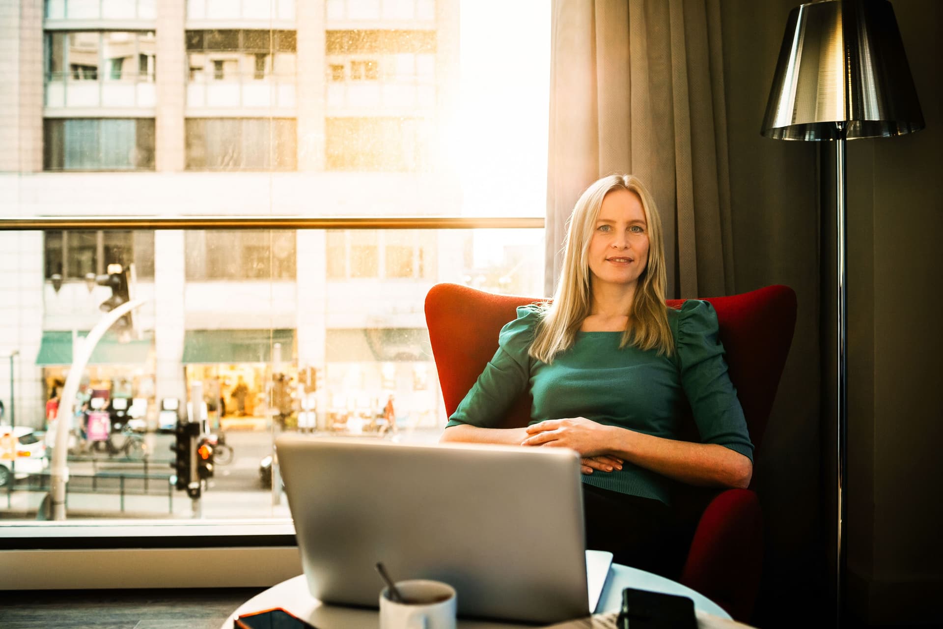 Eine Frau sitzt in einem roten Sessel. Vor ihr steht ein aufgeklappter Laptop. Im Hintergrund ein großes Fenster, durch das die Sonne scheint.