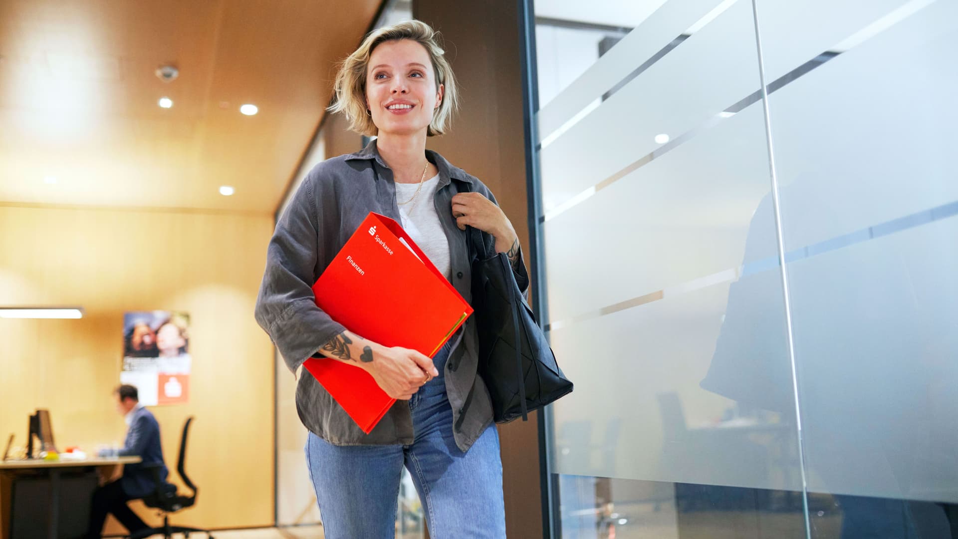 Eine blonde Frau in den 30ern kommt lächelnd von einem Sparkassen-Beratungstermin und hat einen roten Ordner mit dem Aufdruck Finanzen unterm Arm