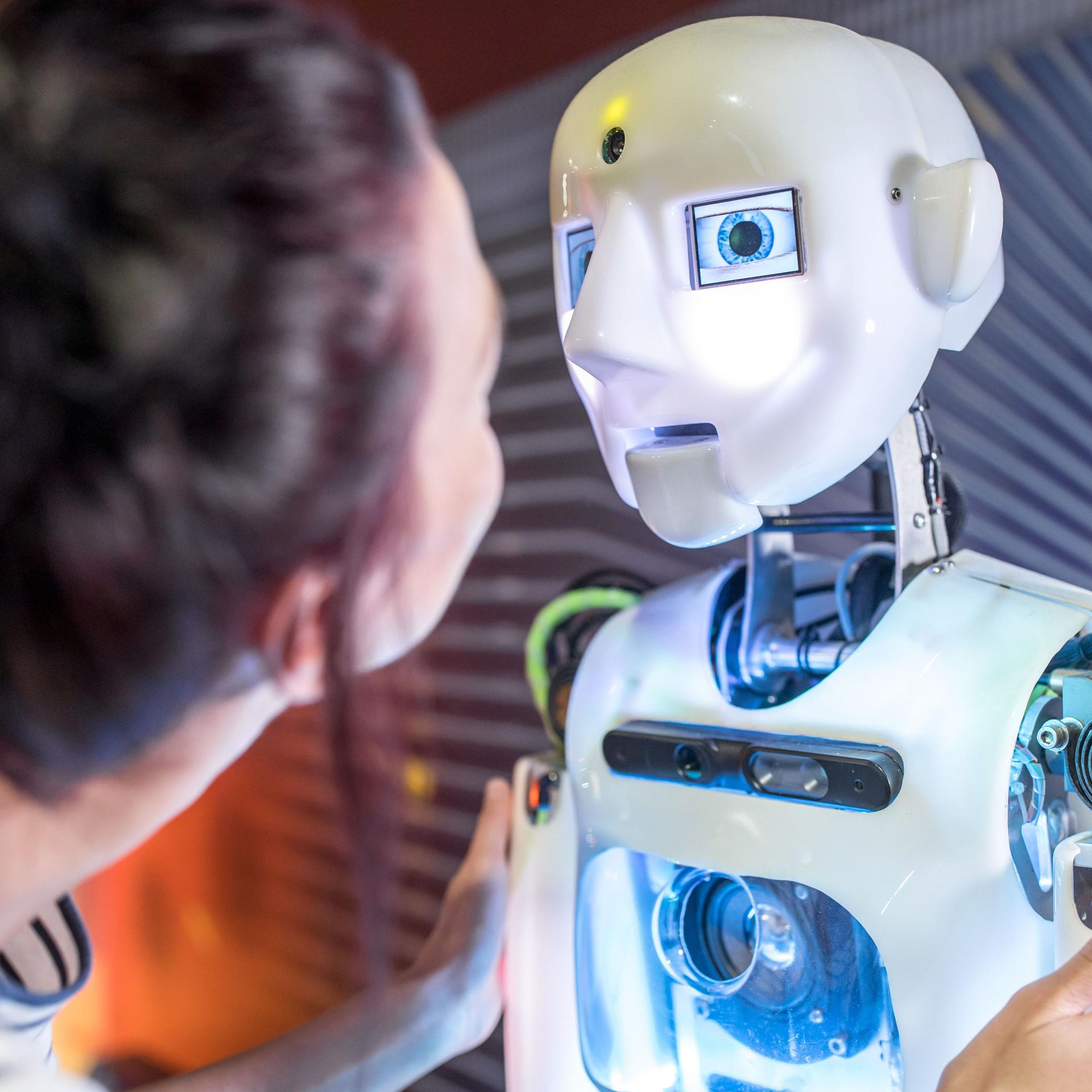 Technische Entwicklerin sieht sich humanoiden Roboter in der Werkstatt an. Er ist weiß und hat neben blauen Augen weitere blau leuchtende Elemente.