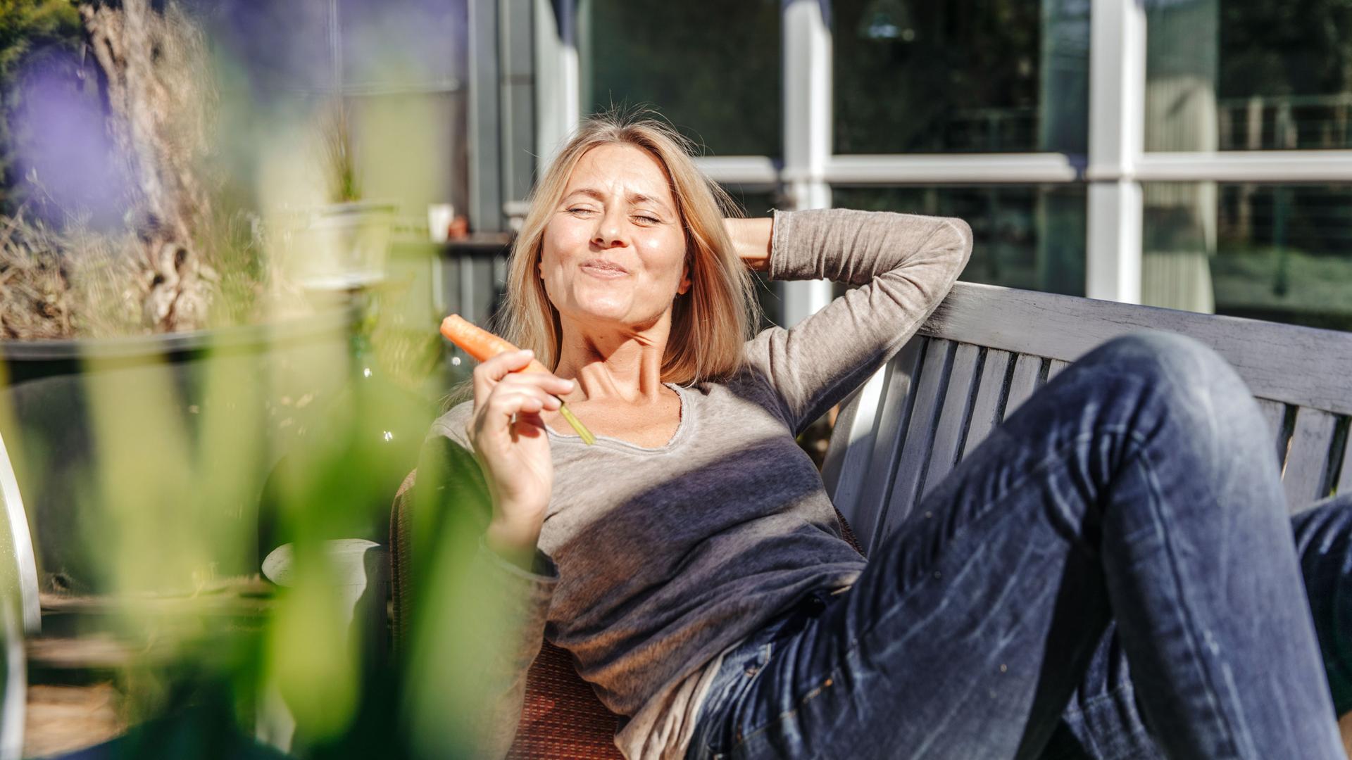Frau entspannt sich auf einer Gartenbank und isst eine Karotte