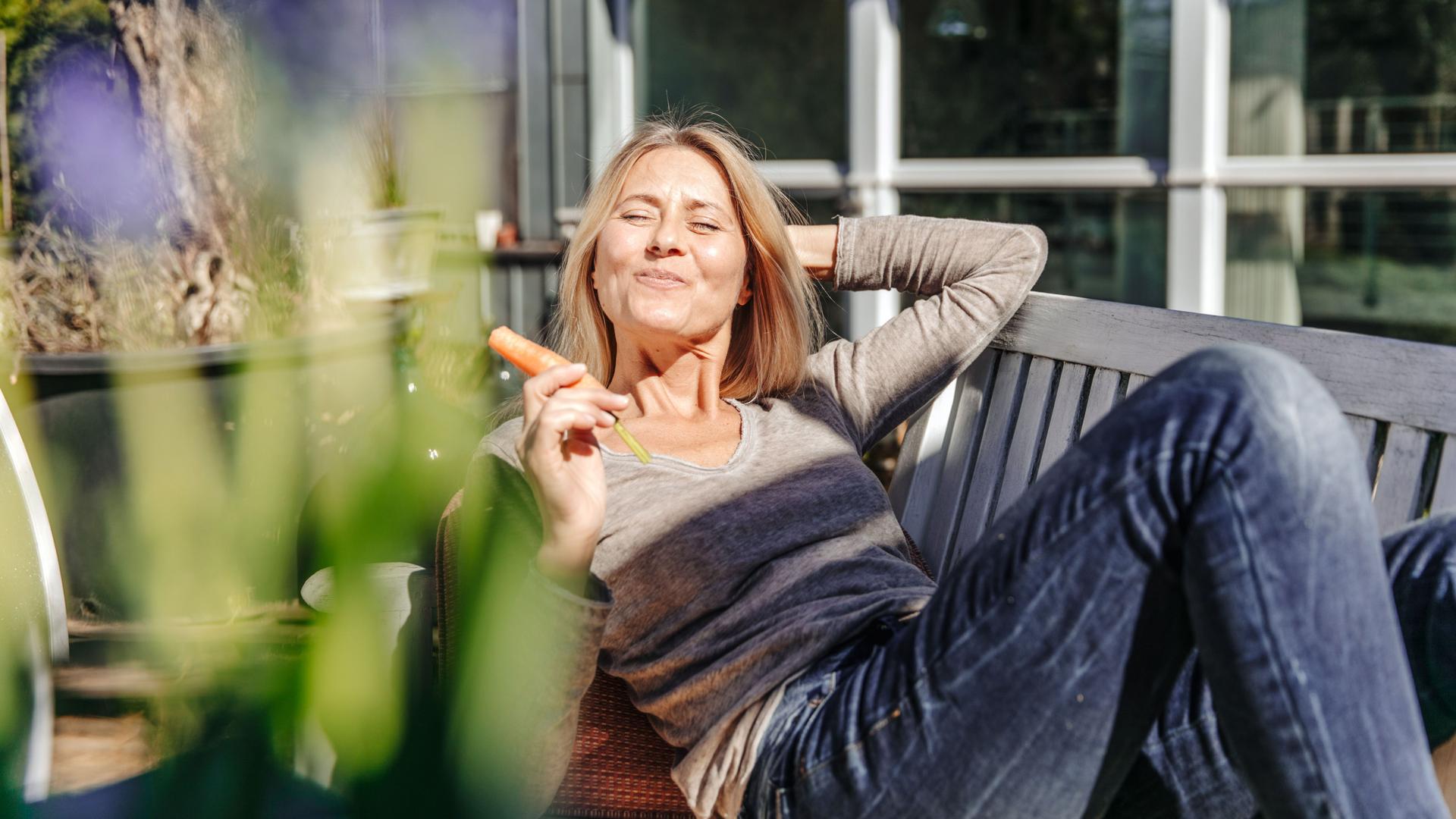 Frau entspannt sich auf einer Gartenbank und isst eine Karotte