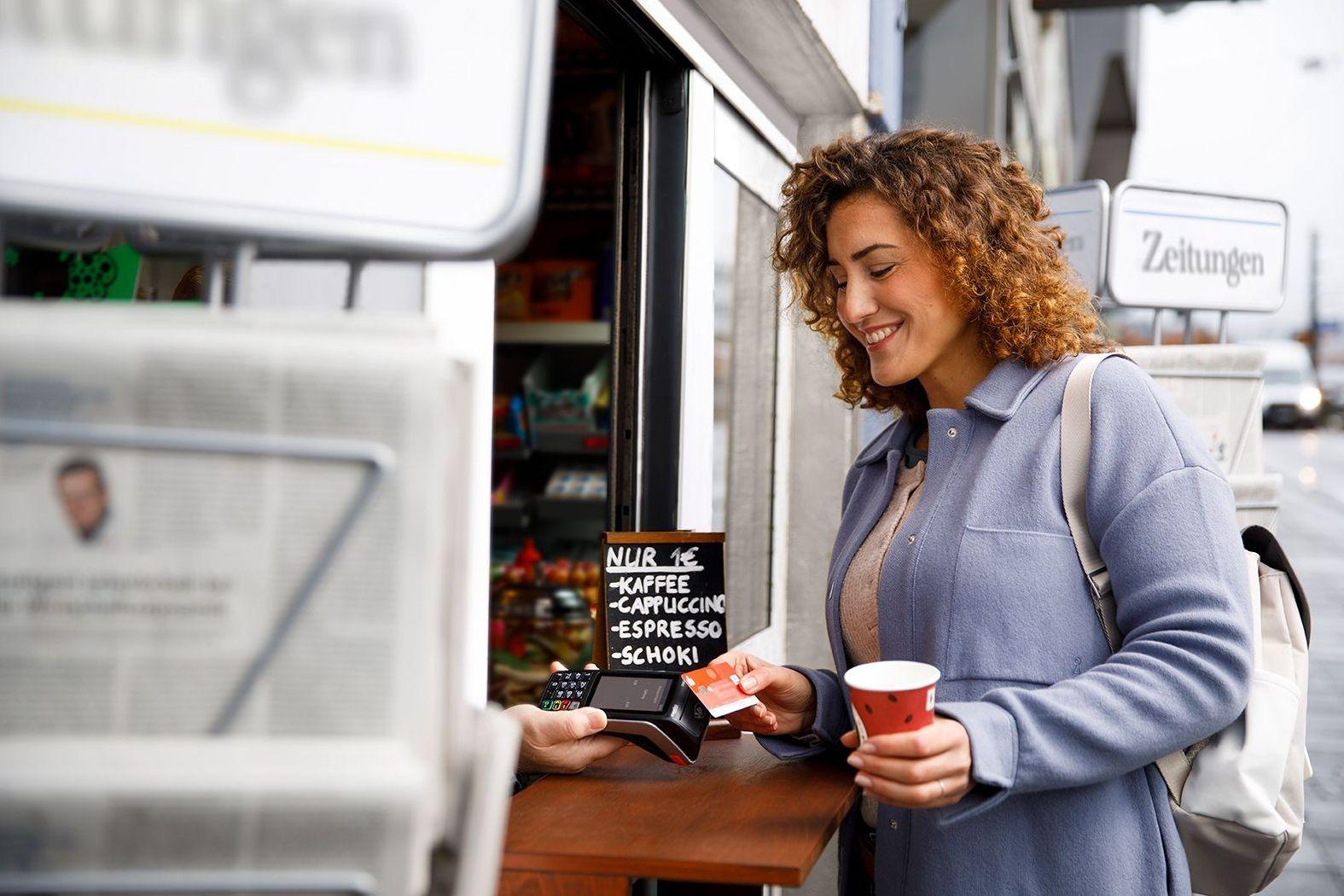 Junge Frau mit dunklen Locken kauft mit der Sparkassenbankkarte ein Heißgetränk an einem Kiosk.