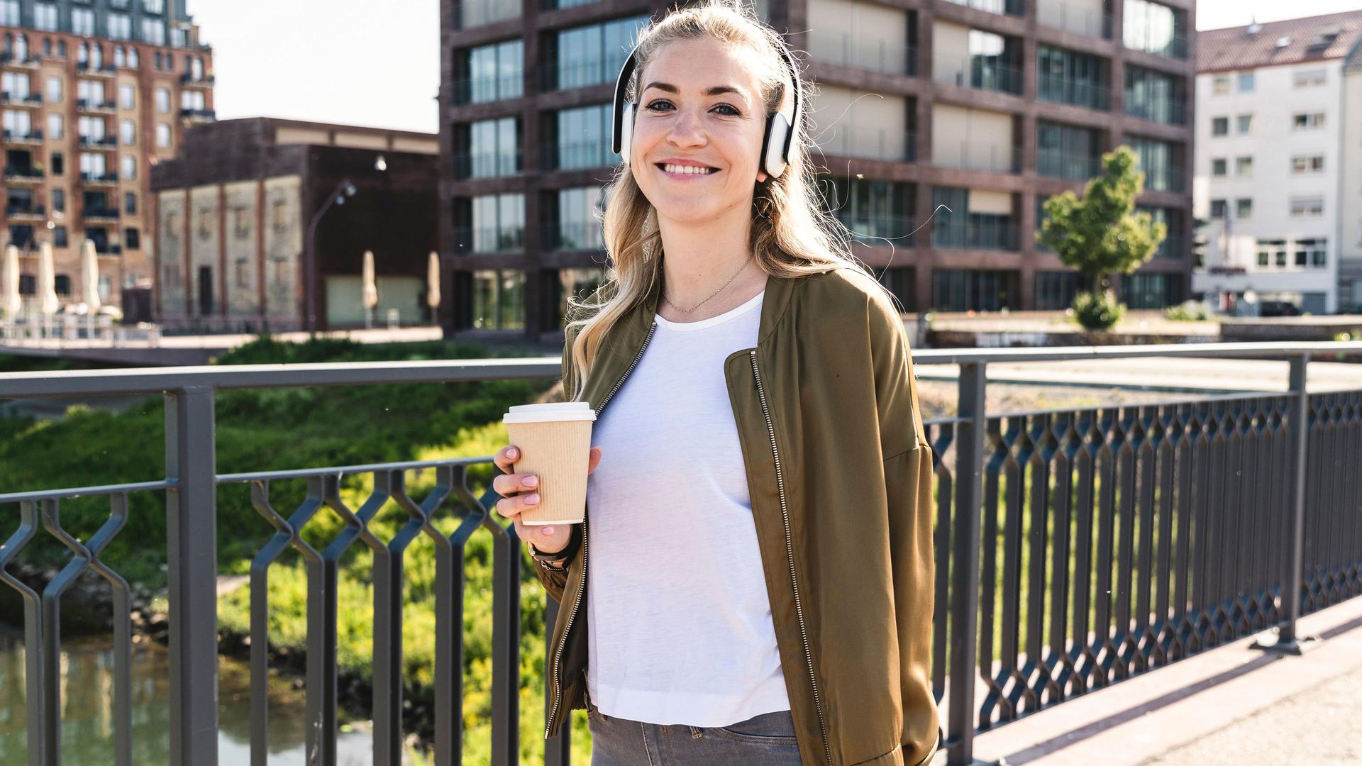 Eine junge Frau spaziert auf einer Brücke mit einem Kaffeebecher. Sie trägt Kopfhörer und lächelt dabei in die Kamera.