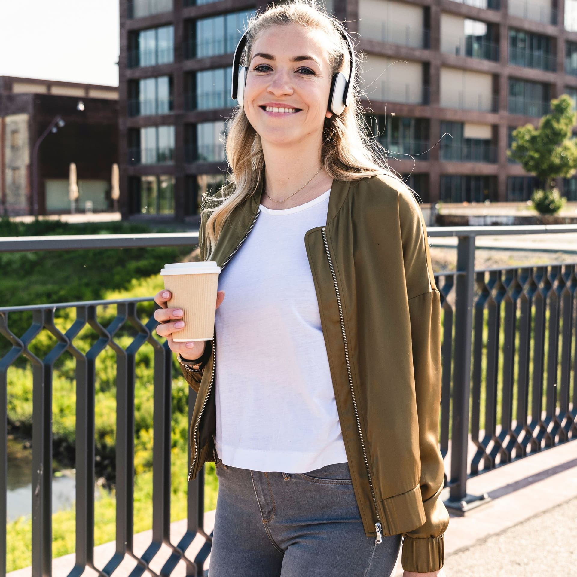 Eine junge Frau spaziert auf einer Brücke mit einem Kaffeebecher. Sie trägt Kopfhörer und lächelt dabei in die Kamera.