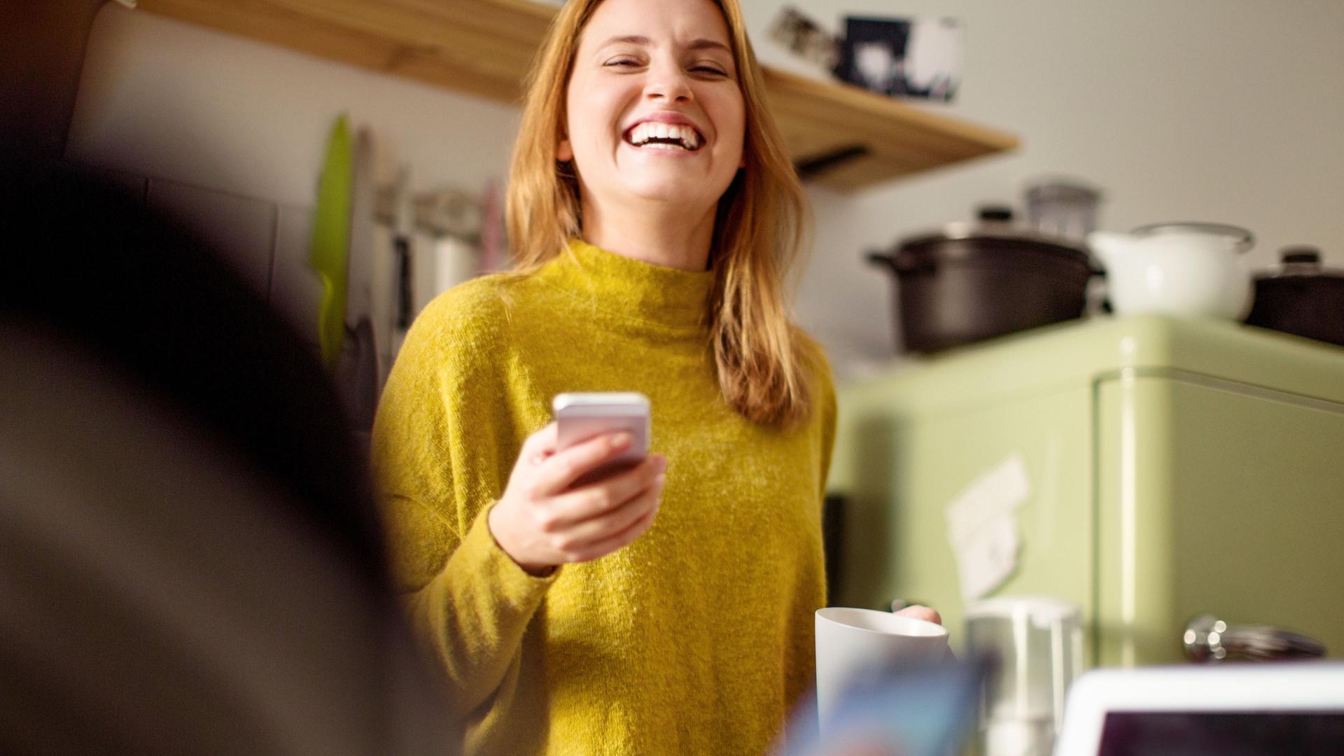 Lachende Frau mit Smartphone in der Hand in der  Küche.