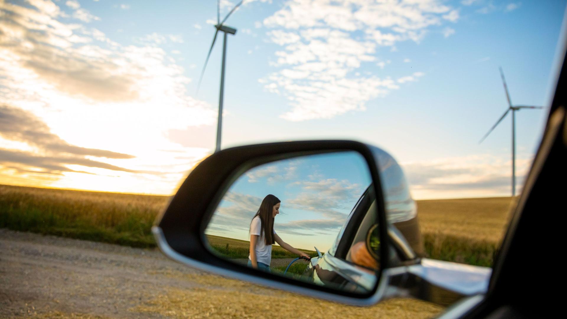 Eine Reflexion im Rückspiegel eines Autos. Darin eine Frau, die ein Elektroauto betankt.