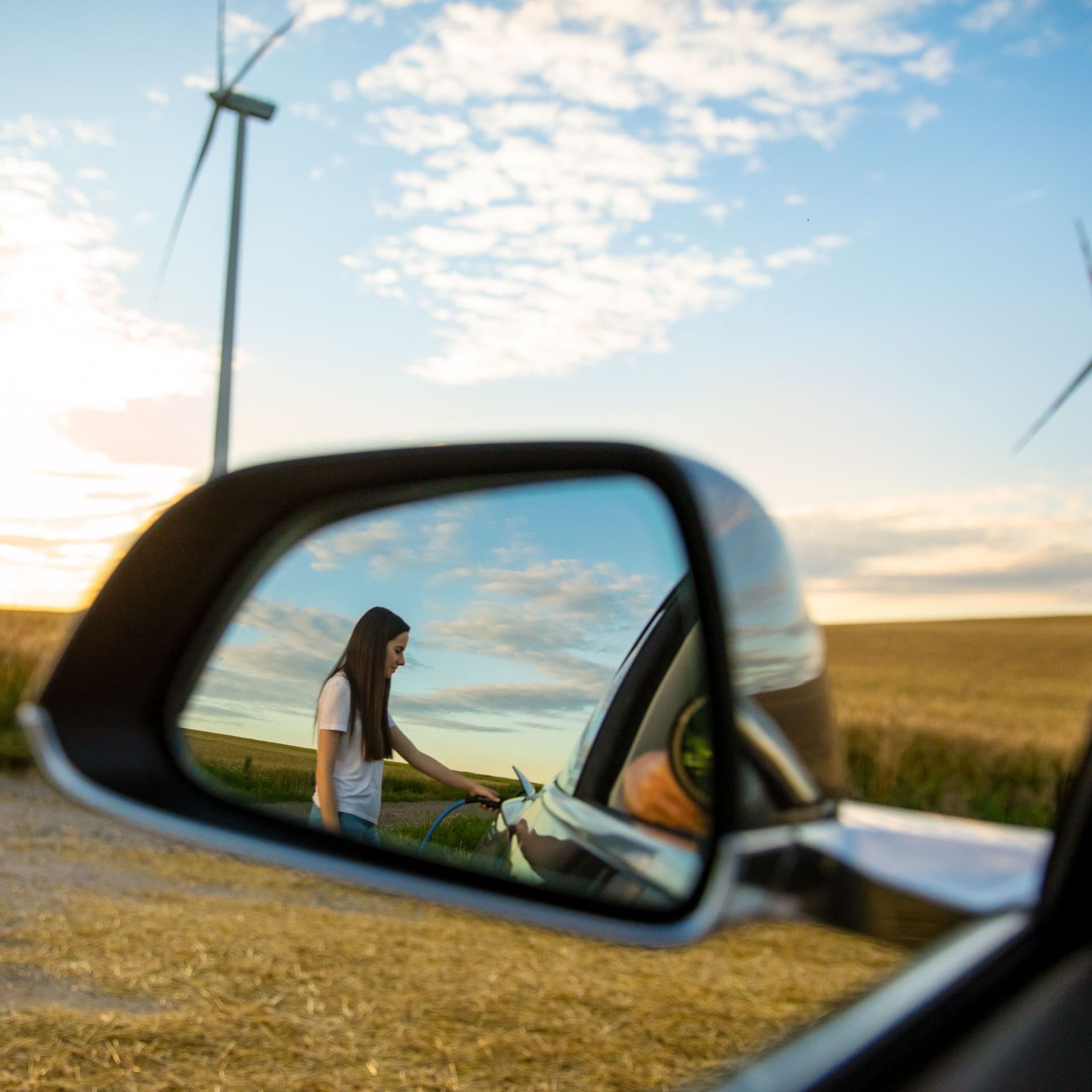Eine Reflexion im Rückspiegel eines Autos. Darin eine Frau, die ein Elektroauto betankt.