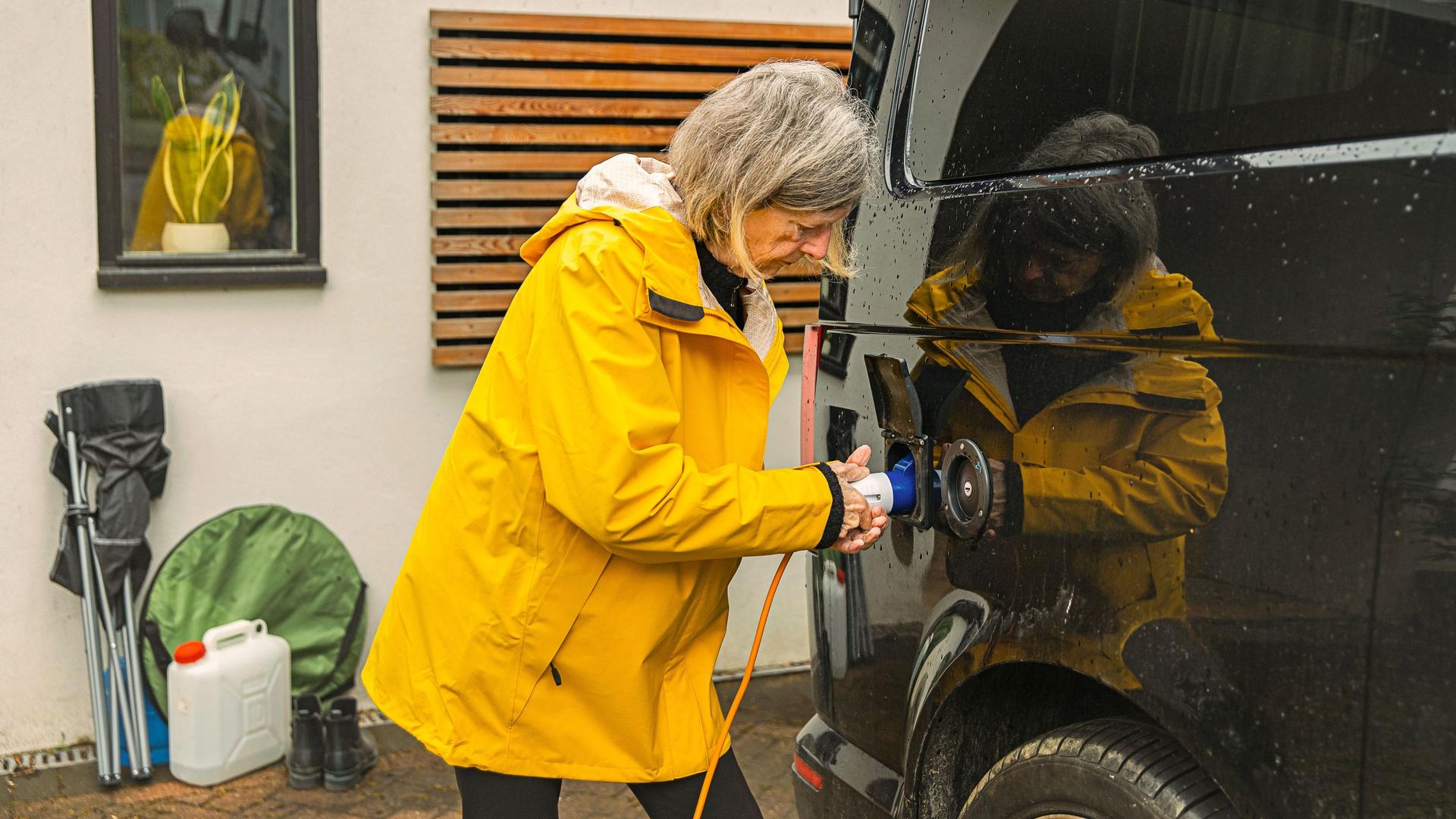 Eine ältere Frau mit gelber Regenjacke lädt ein Elektroauto im Vorgarten auf.