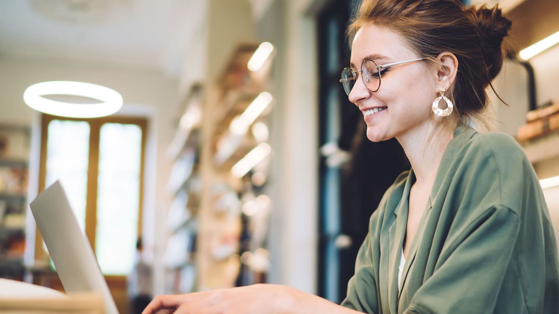 Fröhliche junge Frau schreibt in einer Bibliothek auf ihrem Laptop