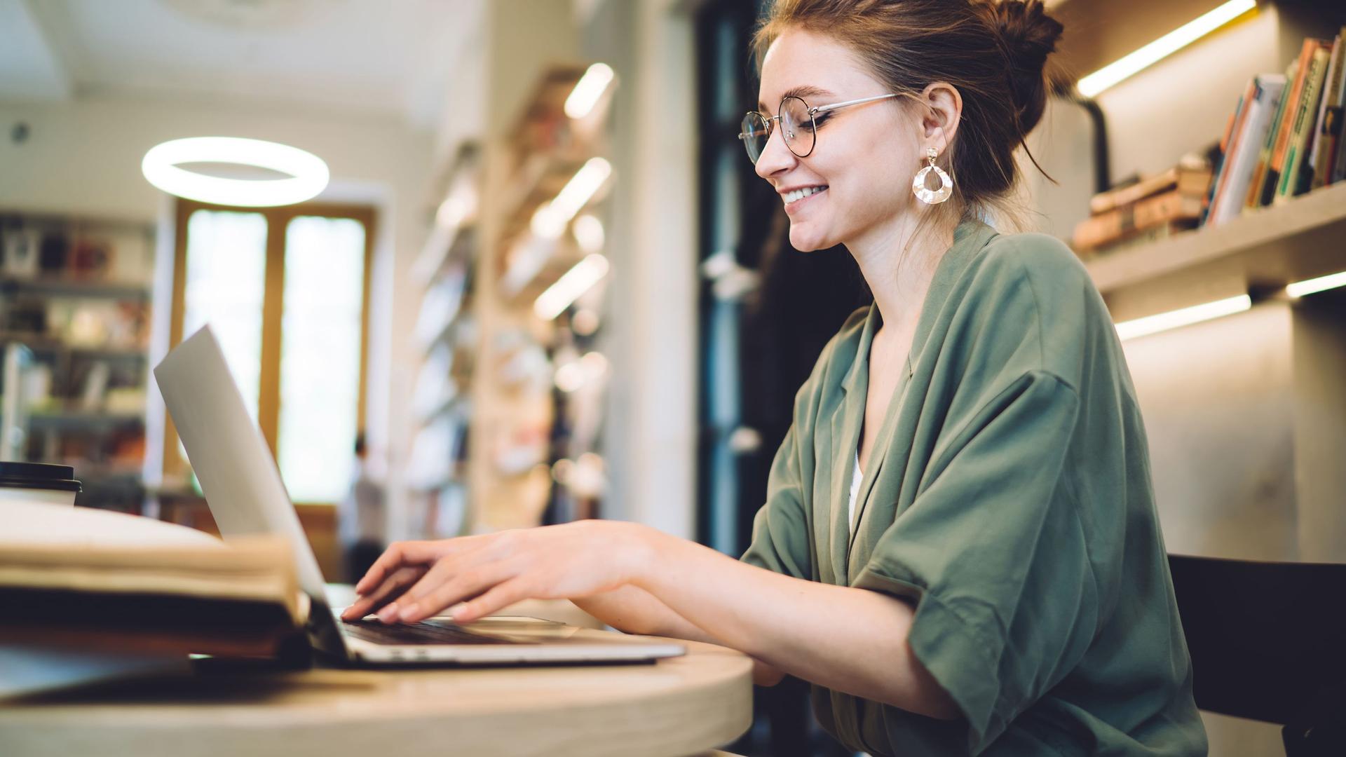 Fröhliche junge Frau schreibt in einer Bibliothek auf ihrem Laptop