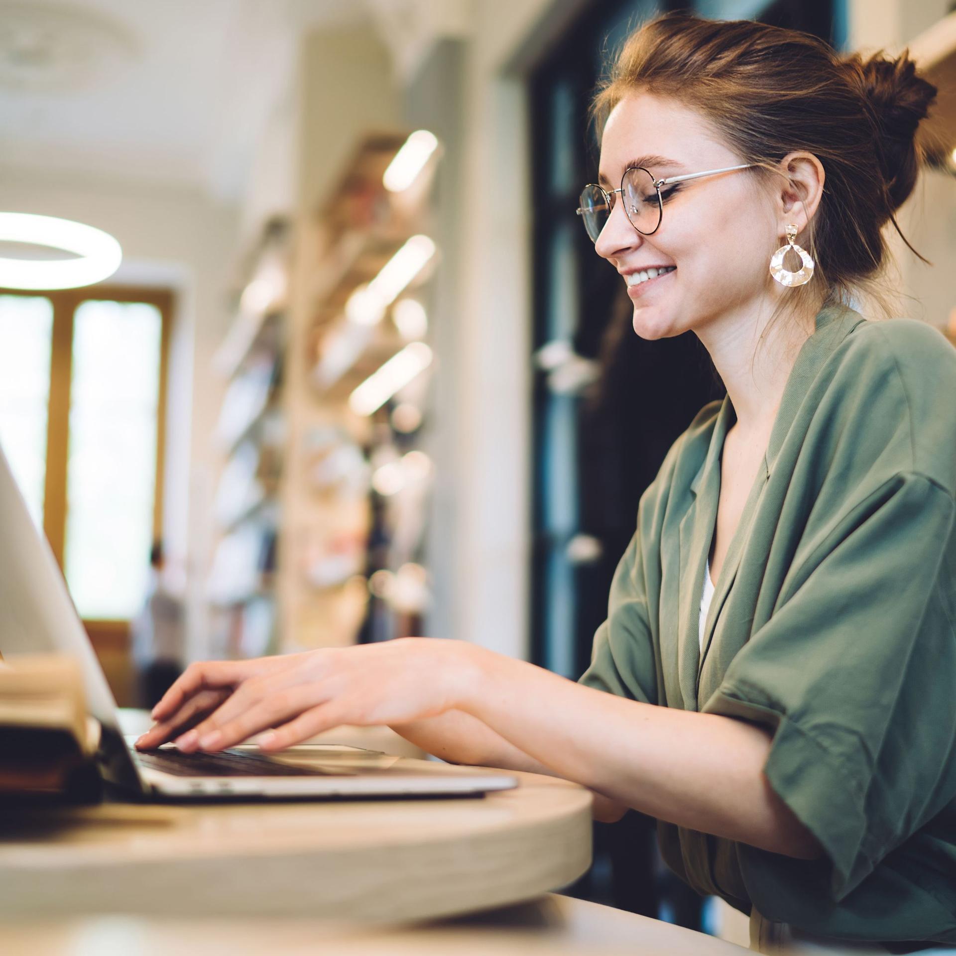 Fröhliche junge Frau schreibt in einer Bibliothek auf ihrem Laptop