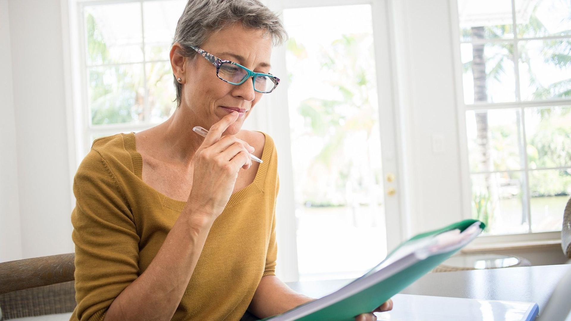 Eine ältere Frau mit Brille, leicht grauen Haaren und ockerfarbenem Pulli sitzt an einem Tisch in einer modernen Wohnung. Sie studiert konzentriert Unterlagen in ihrer Hand.